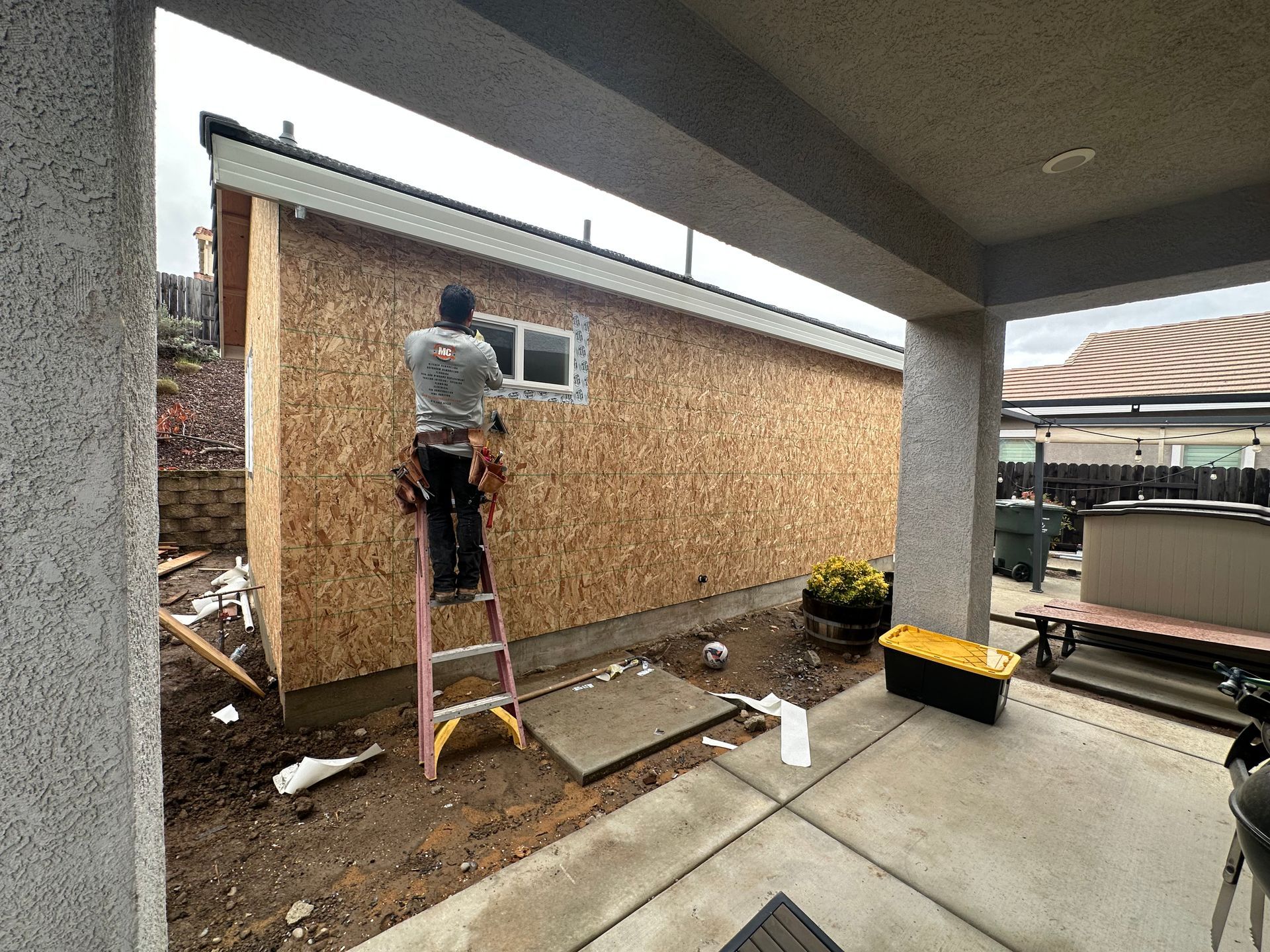 Person on a ladder repairing a wall beside a covered patio.