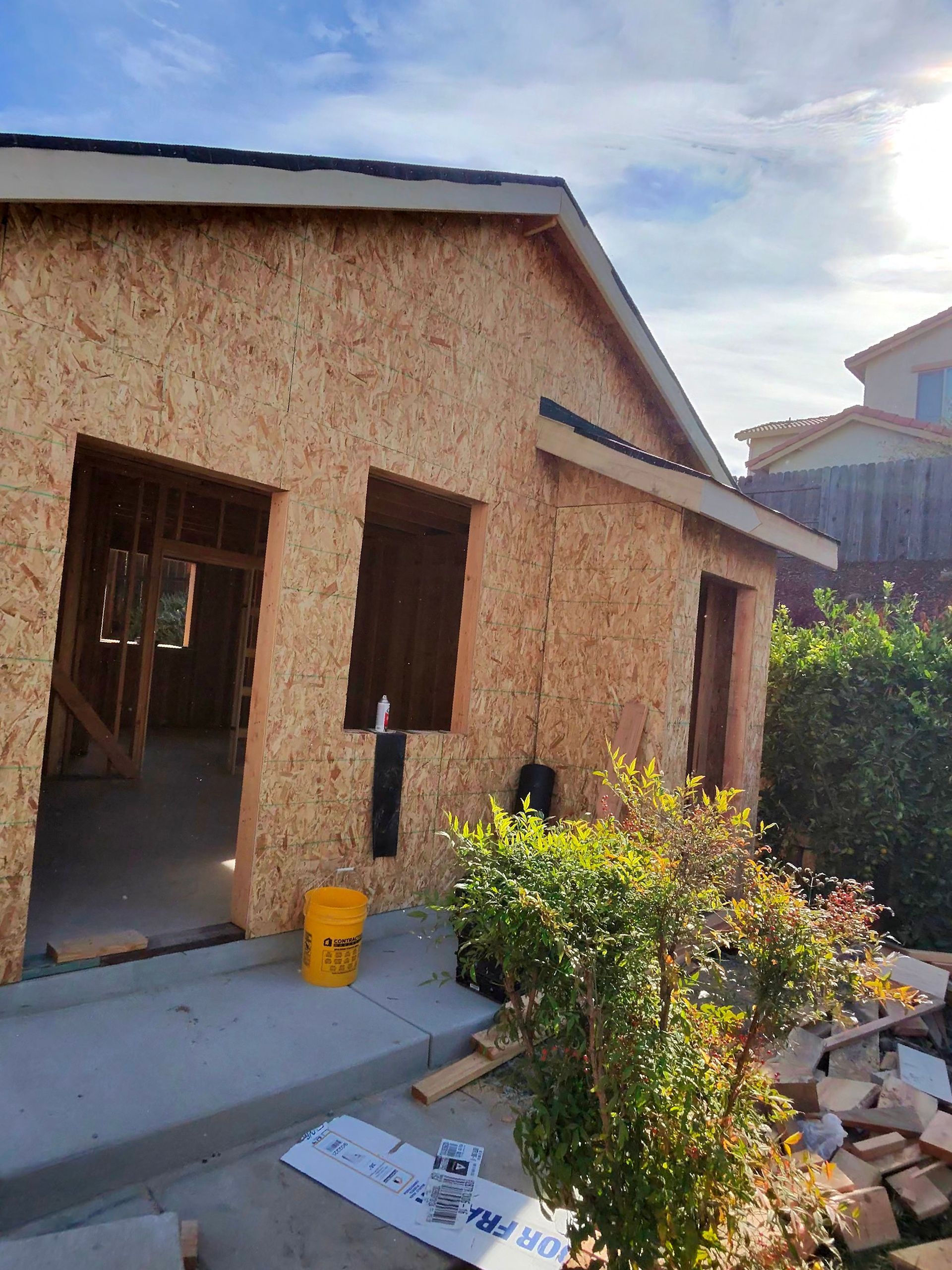 Small unfinished house exterior with open door and windows, concrete patio, and shrubs in front.