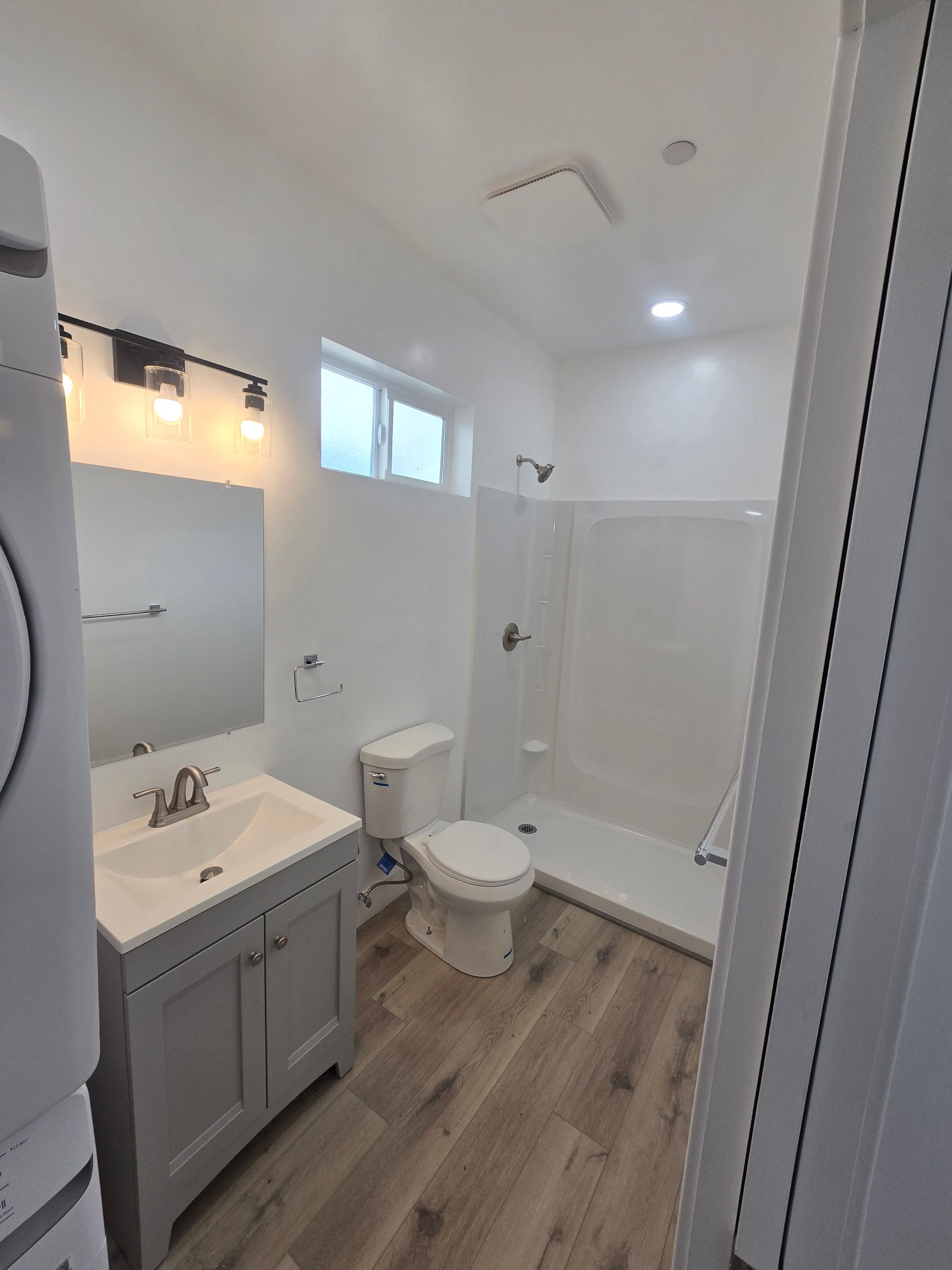 Bright bathroom with white shower, gray vanity, toilet, and wood-look floor, viewed from the doorway