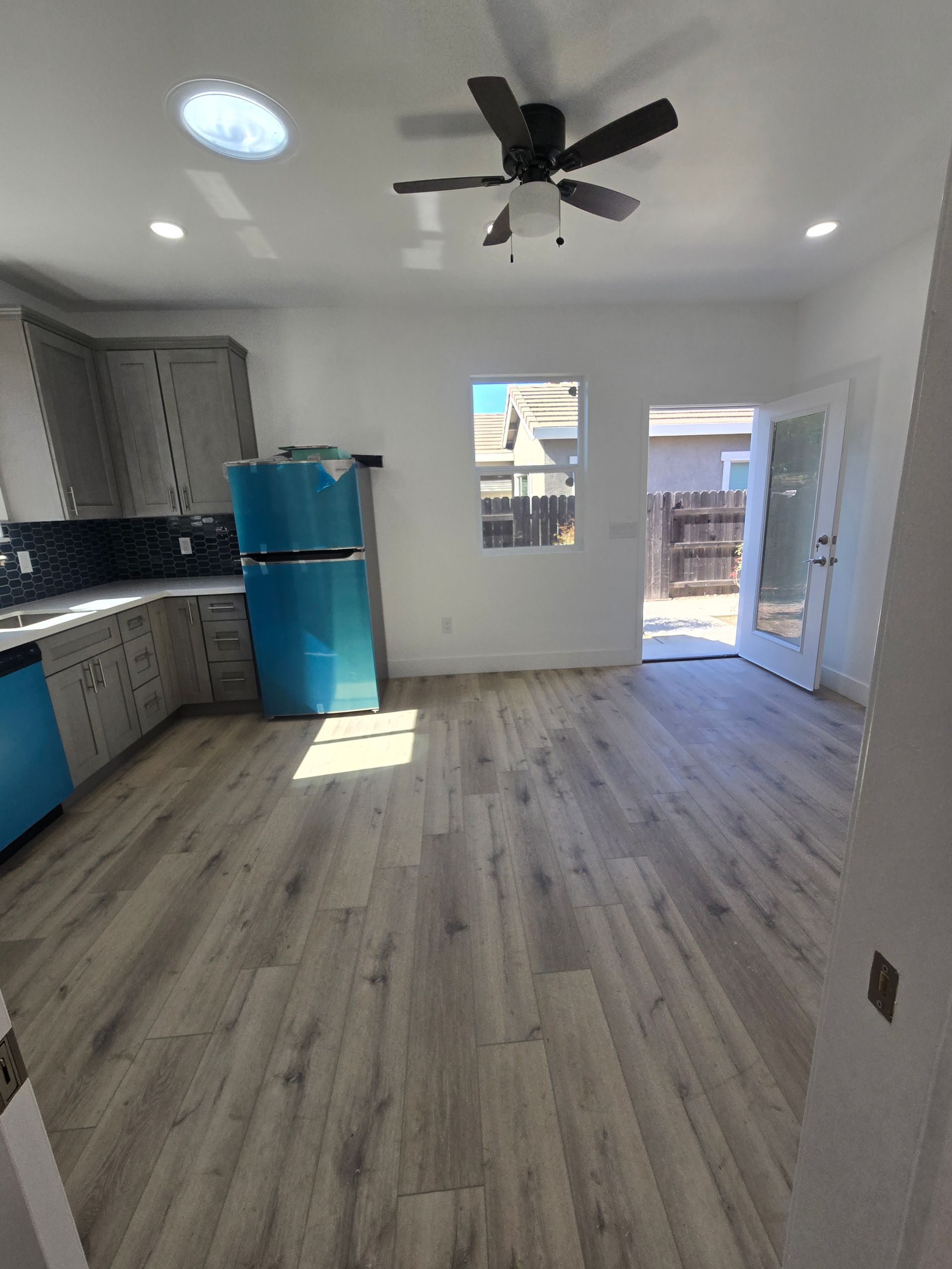 Empty modern kitchen with gray cabinets, blue fridge, wood floor, ceiling fan, and open door to outside.