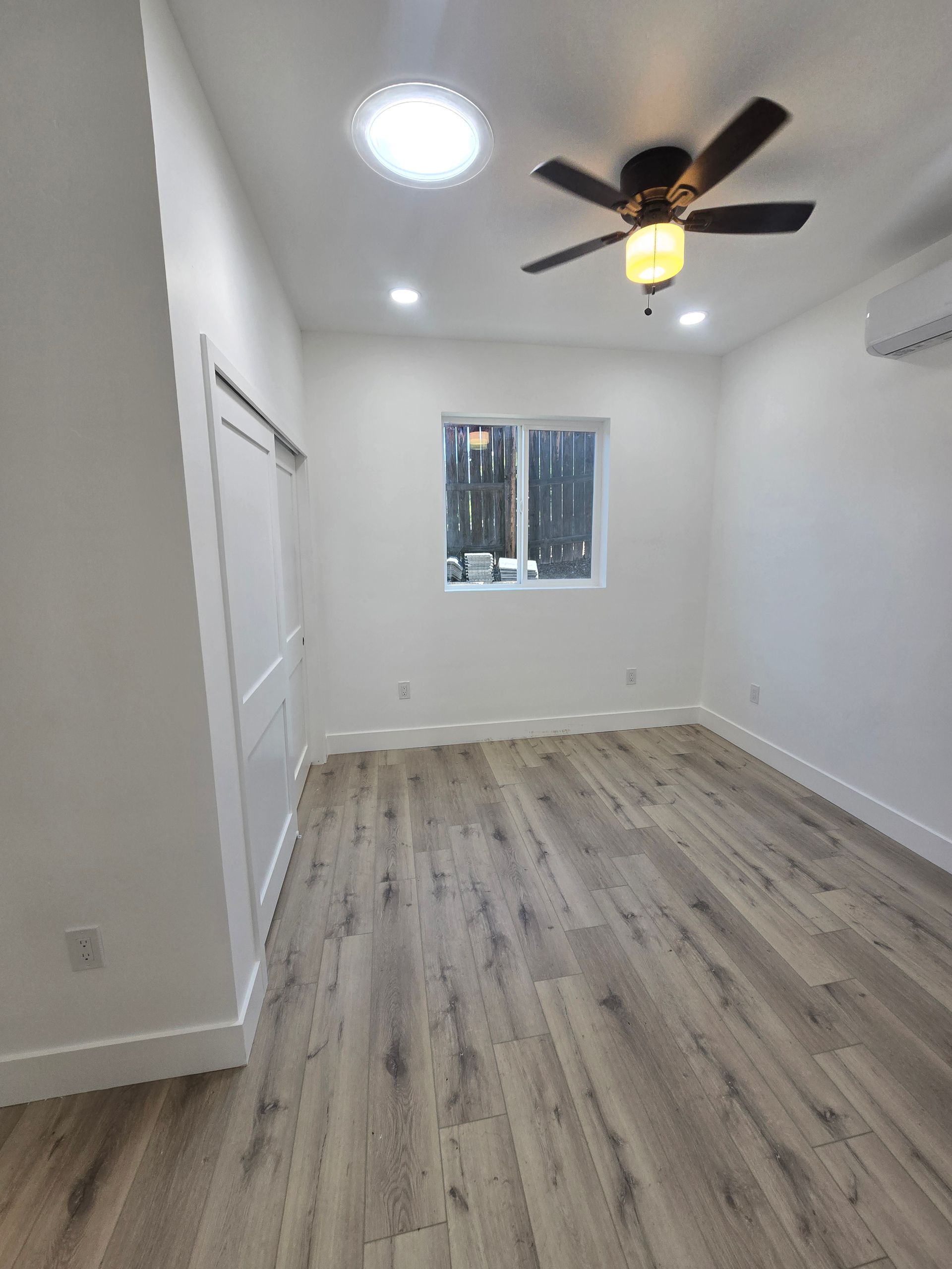 Empty white bedroom with wood flooring, ceiling fan, recessed light, and a small window.