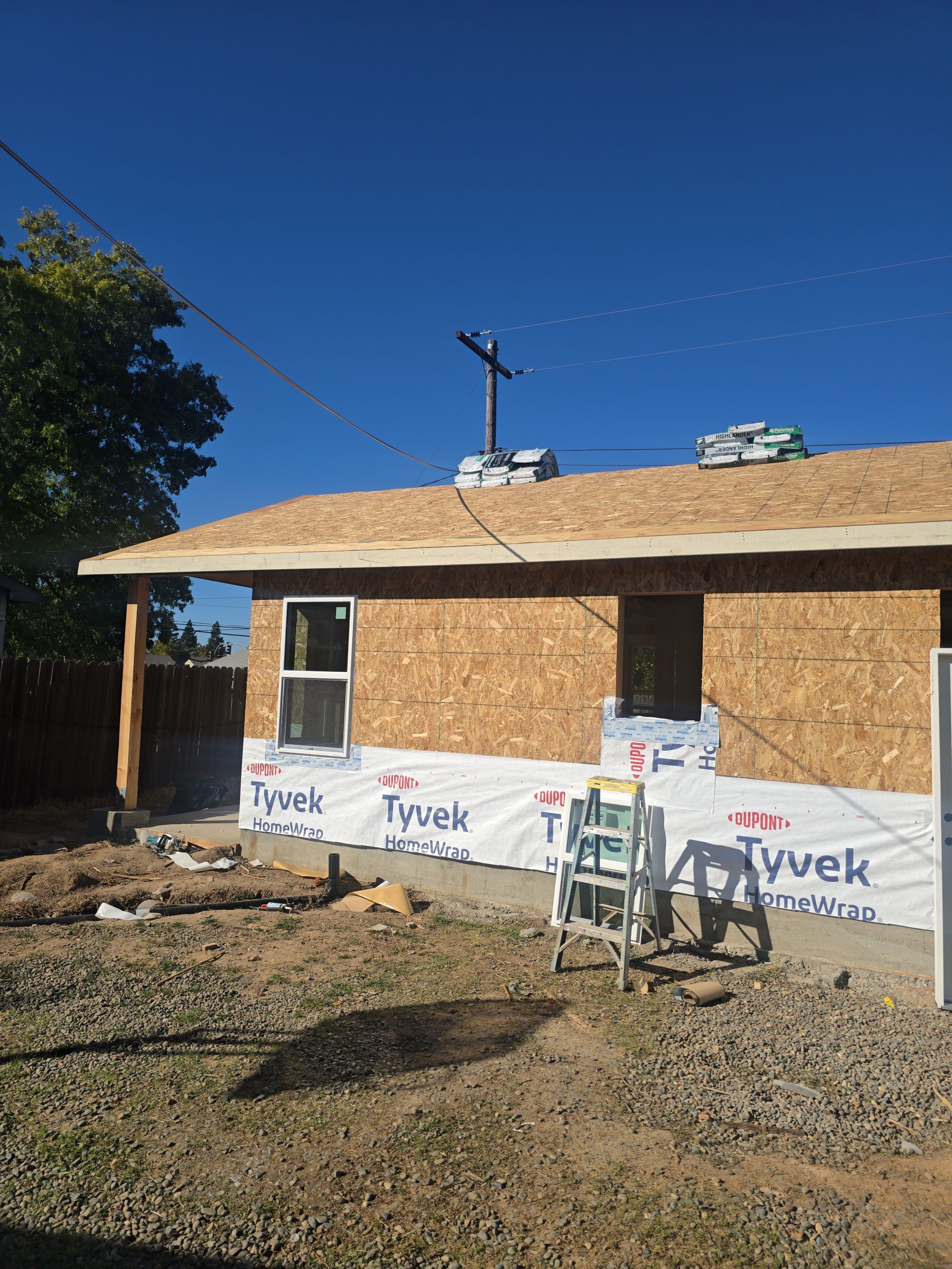 House under construction with roof sheathing, Tyvek wrap, and a gravel yard under a clear blue sky
