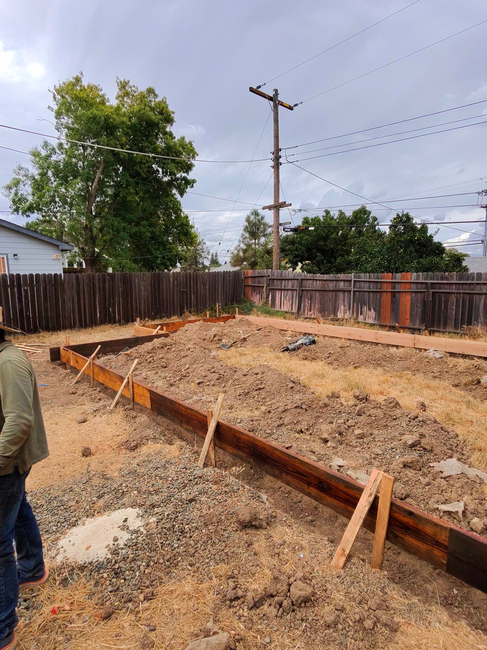 Backyard construction site with wooden forms, gravel piles, fence, and utility pole under cloudy sky