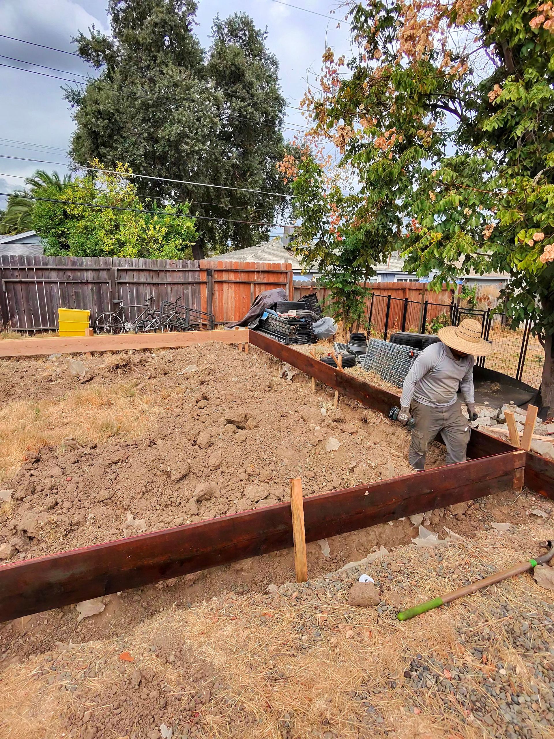 Backyard construction area with wooden formwork, dry ground, tools, and a worker near a fence and trees