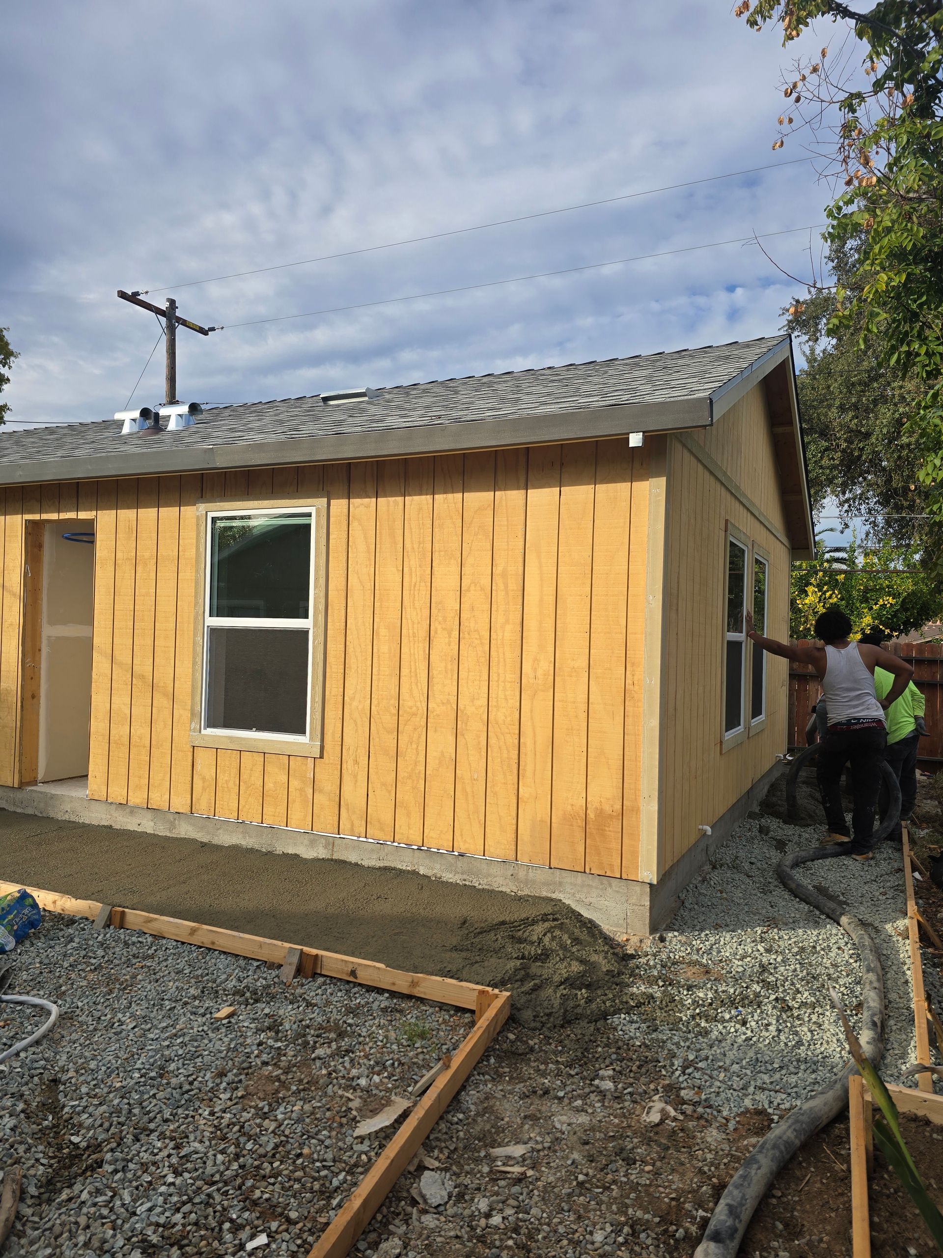 Small tan house with white-trimmed windows on gravel, with workers beside it and a cloudy sky overhead.