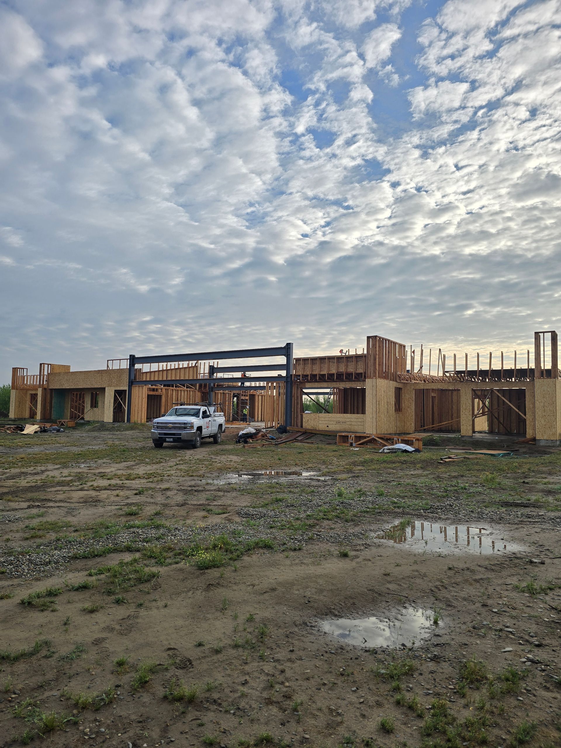 Partly built wooden structure on a muddy lot under a cloudy sky, with a white van nearby