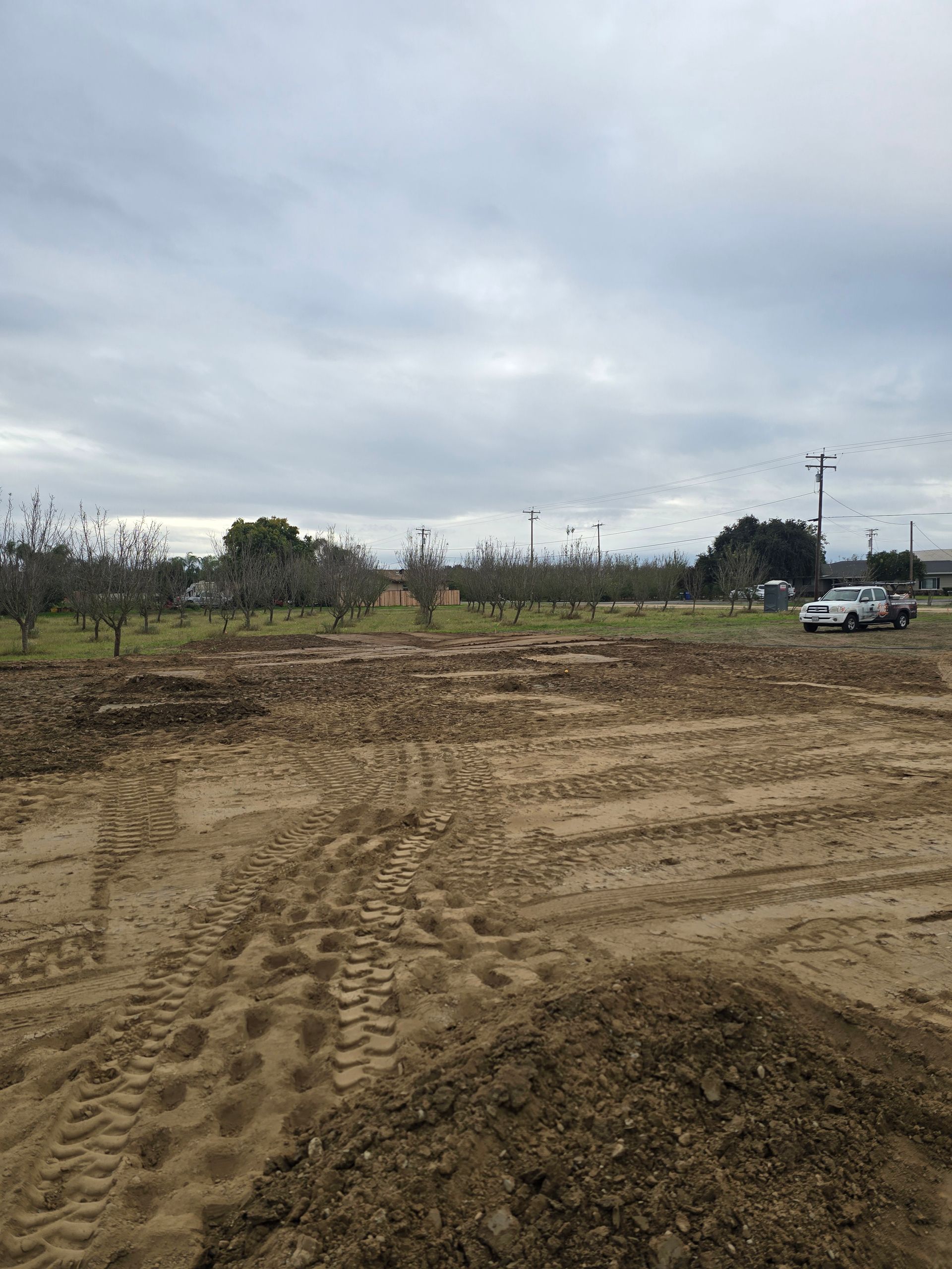 Muddy dirt lot under a cloudy sky, with tire tracks and a car near trees on the right