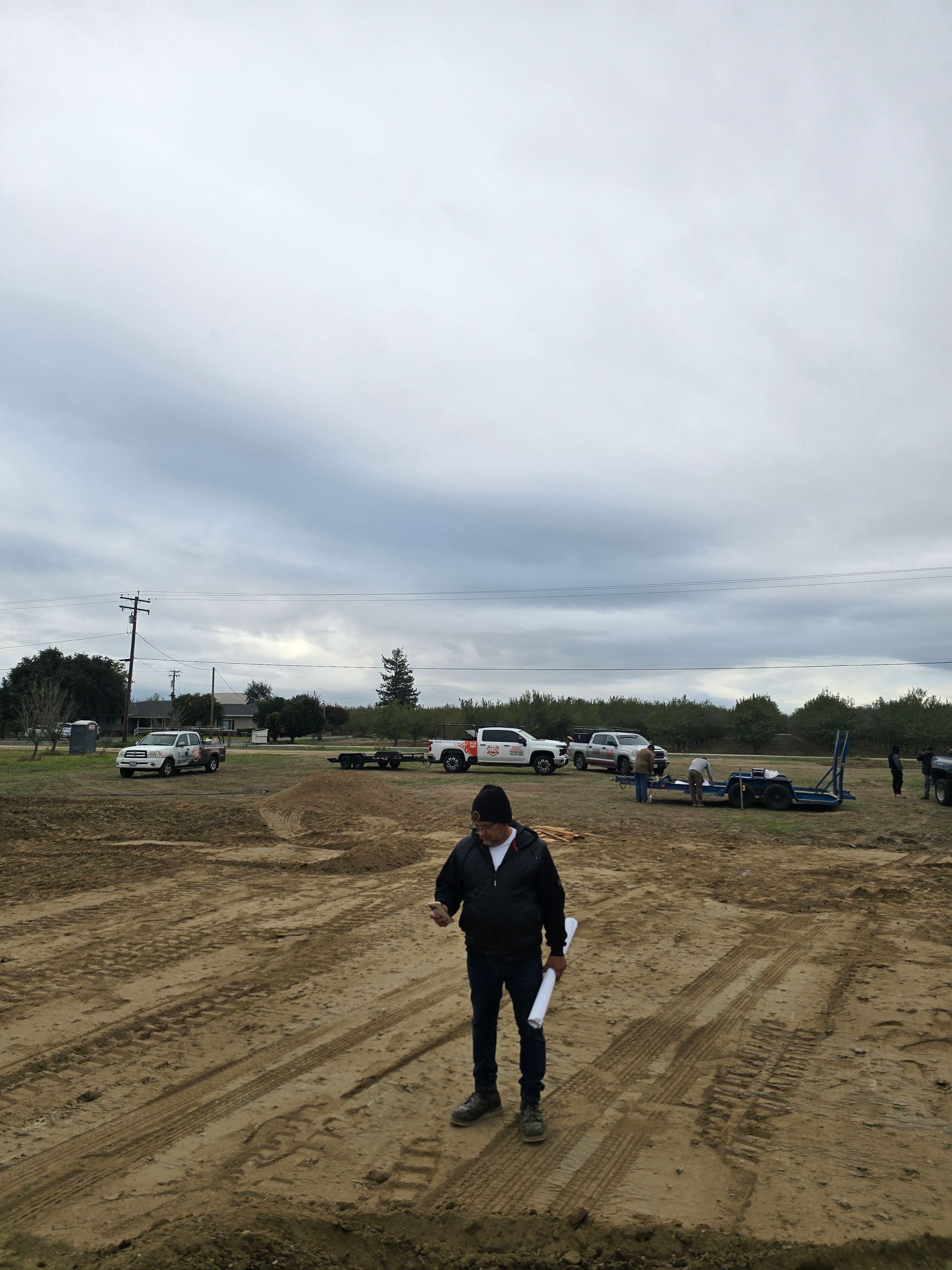 Person standing in a muddy open field with cars and trees under a cloudy sky.
