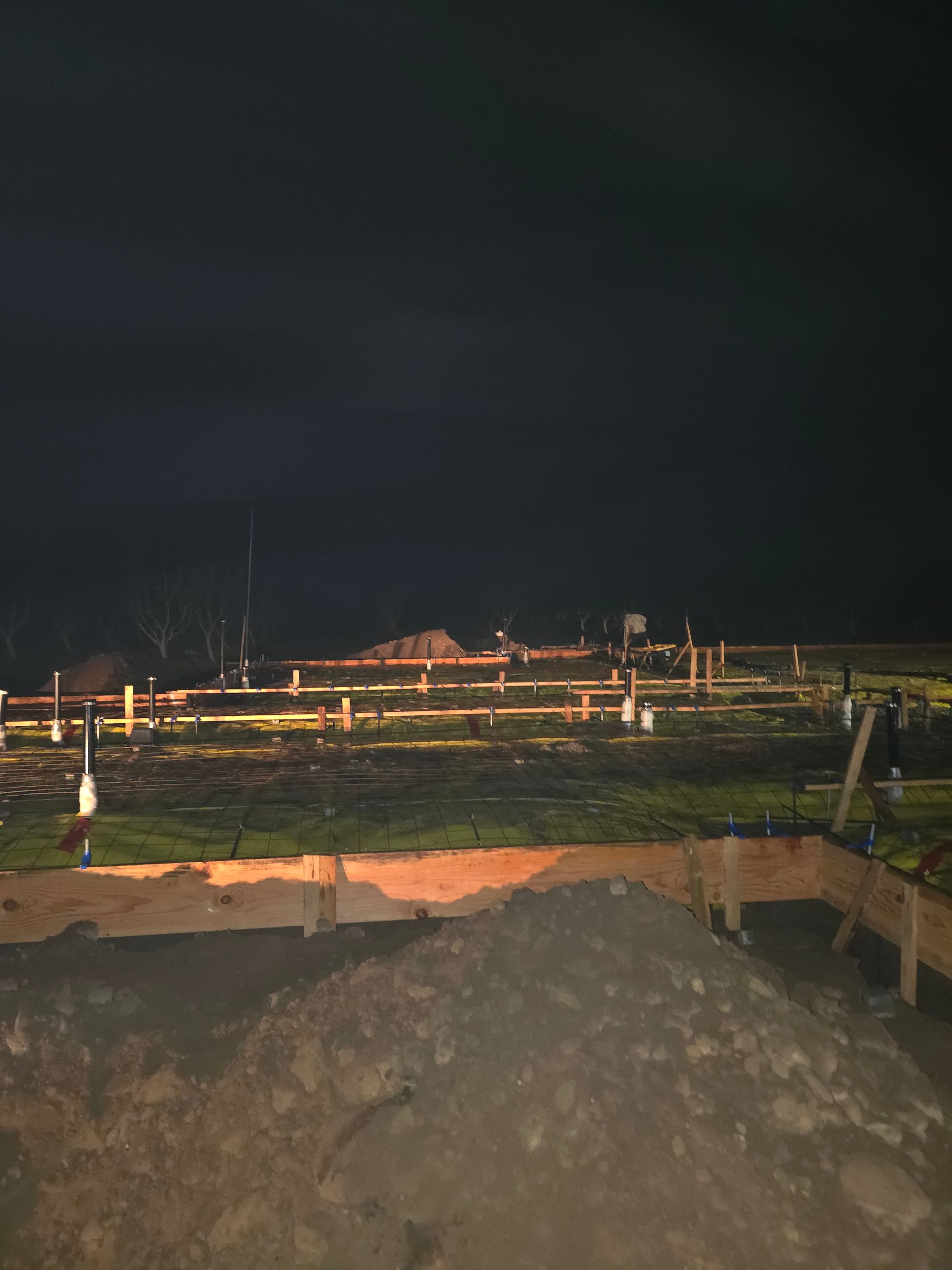 Nighttime construction site with wooden forms, dirt piles, and dim lights under a dark sky