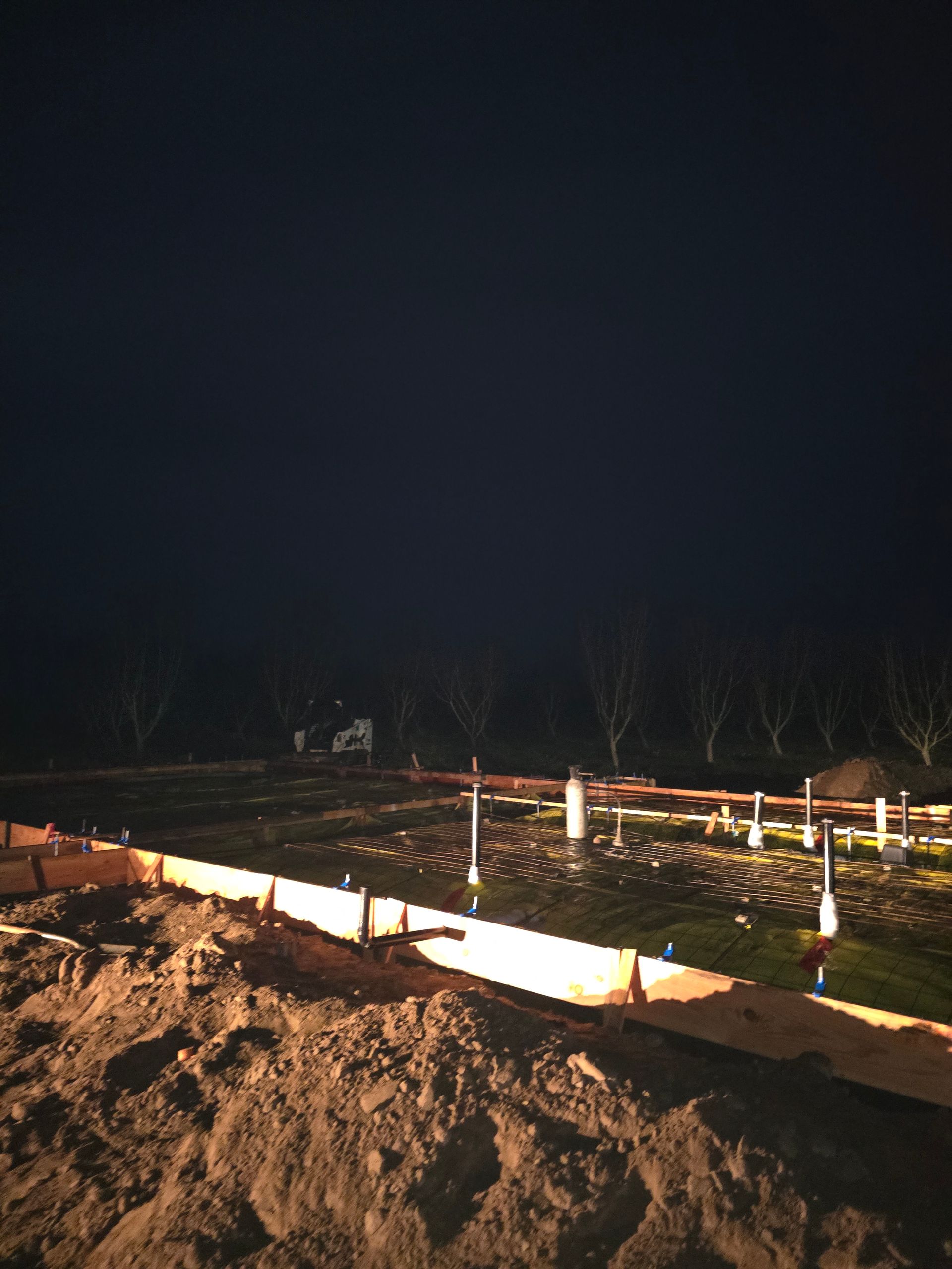 Nighttime construction site with a dirt trench, orange-lit boards, and white stakes marking the ground