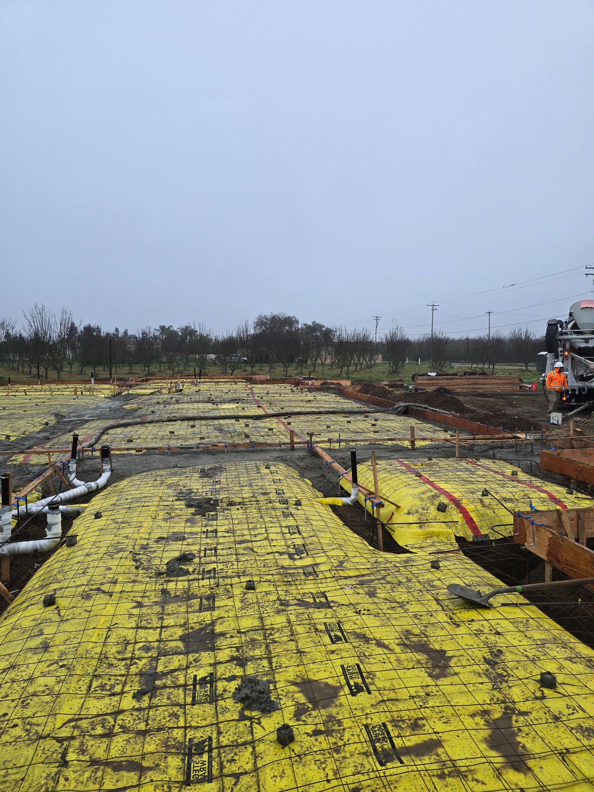 Construction site with yellow formwork and rebar under a cloudy sky, workers visible in the distance