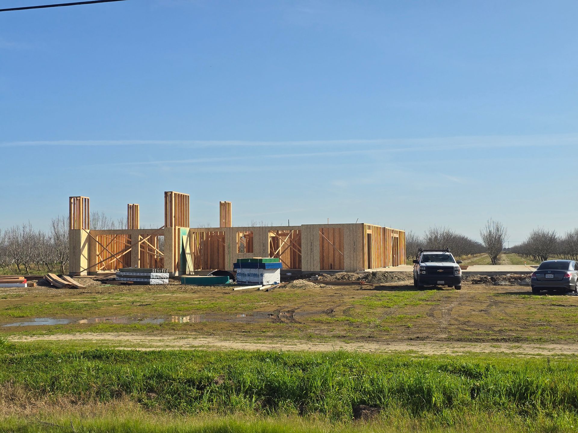 Partially built wooden structure on a grassy field with several parked vehicles under a clear blue sky