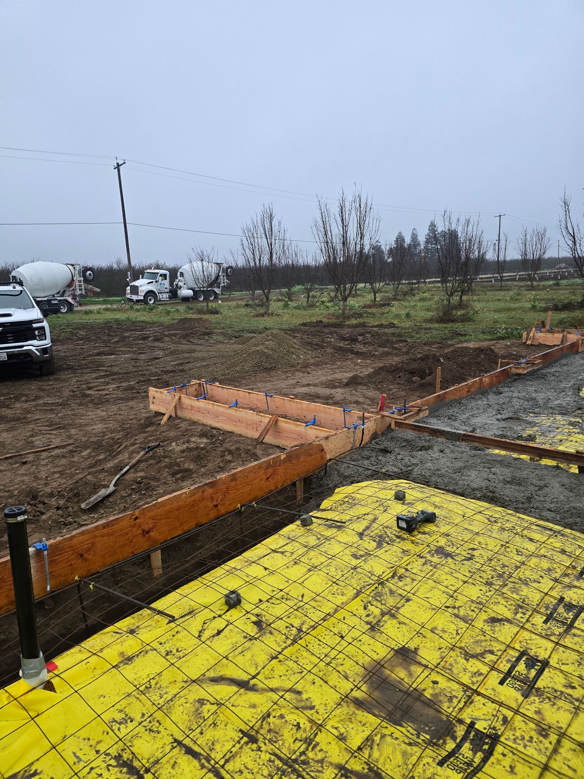 Rainy outdoor lot with yellow paving, orange trench, and parked vehicles under a gray sky