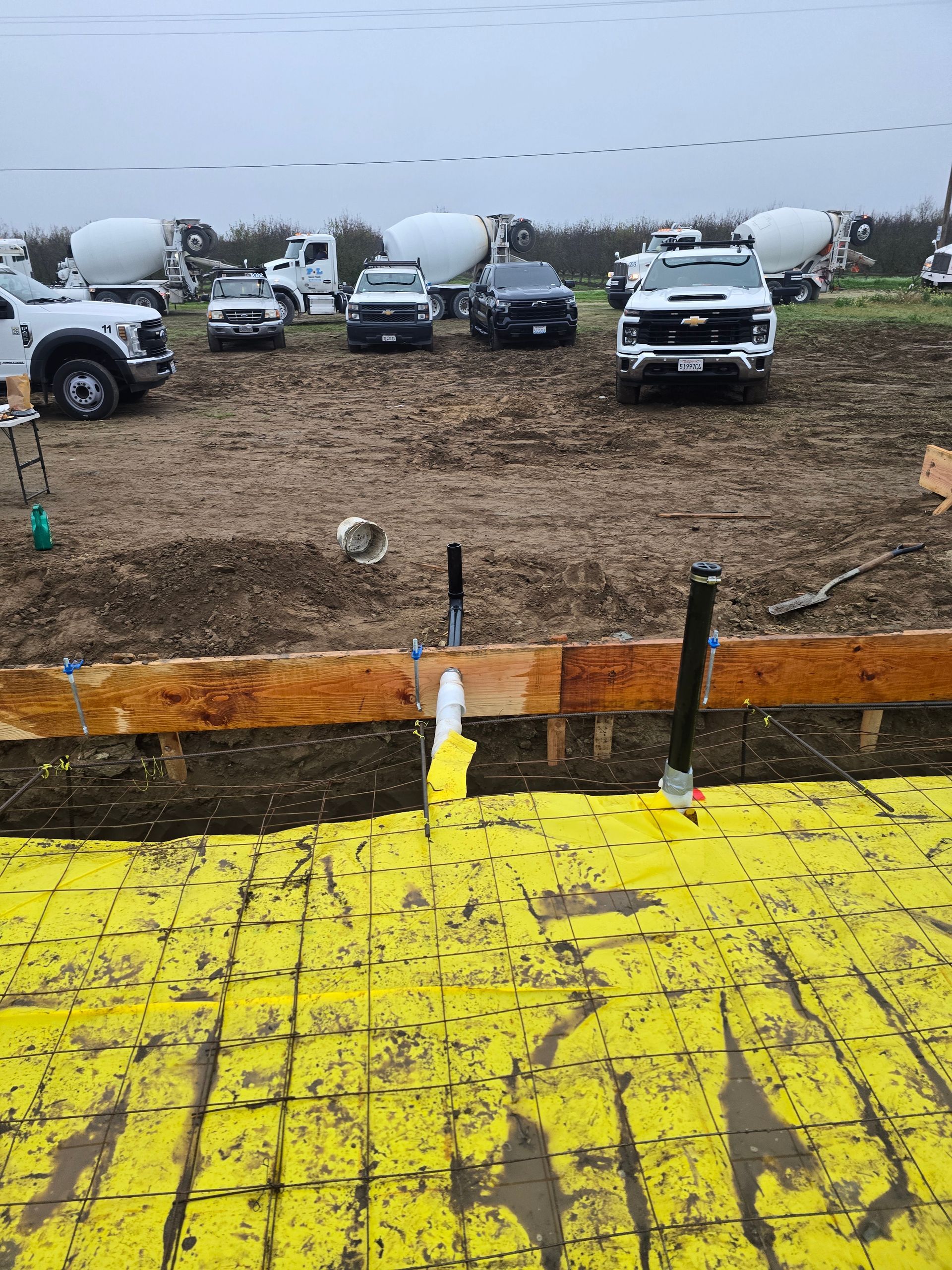 Rainy gravel lot with parked trucks and cement mixers behind a yellow wooden barrier