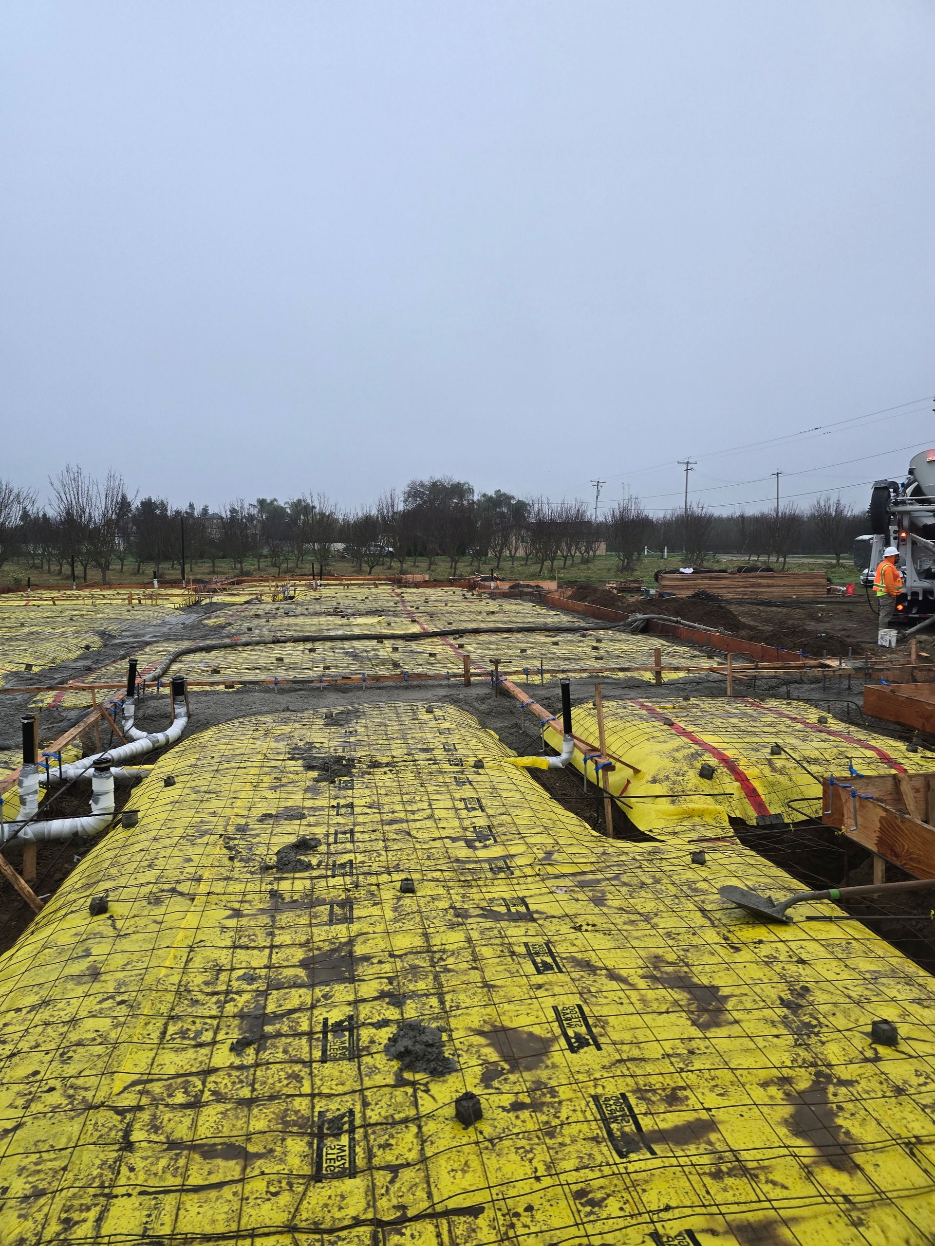 Yellow waterproof membrane laid over a foundation site under a gray sky, with construction machinery at right.