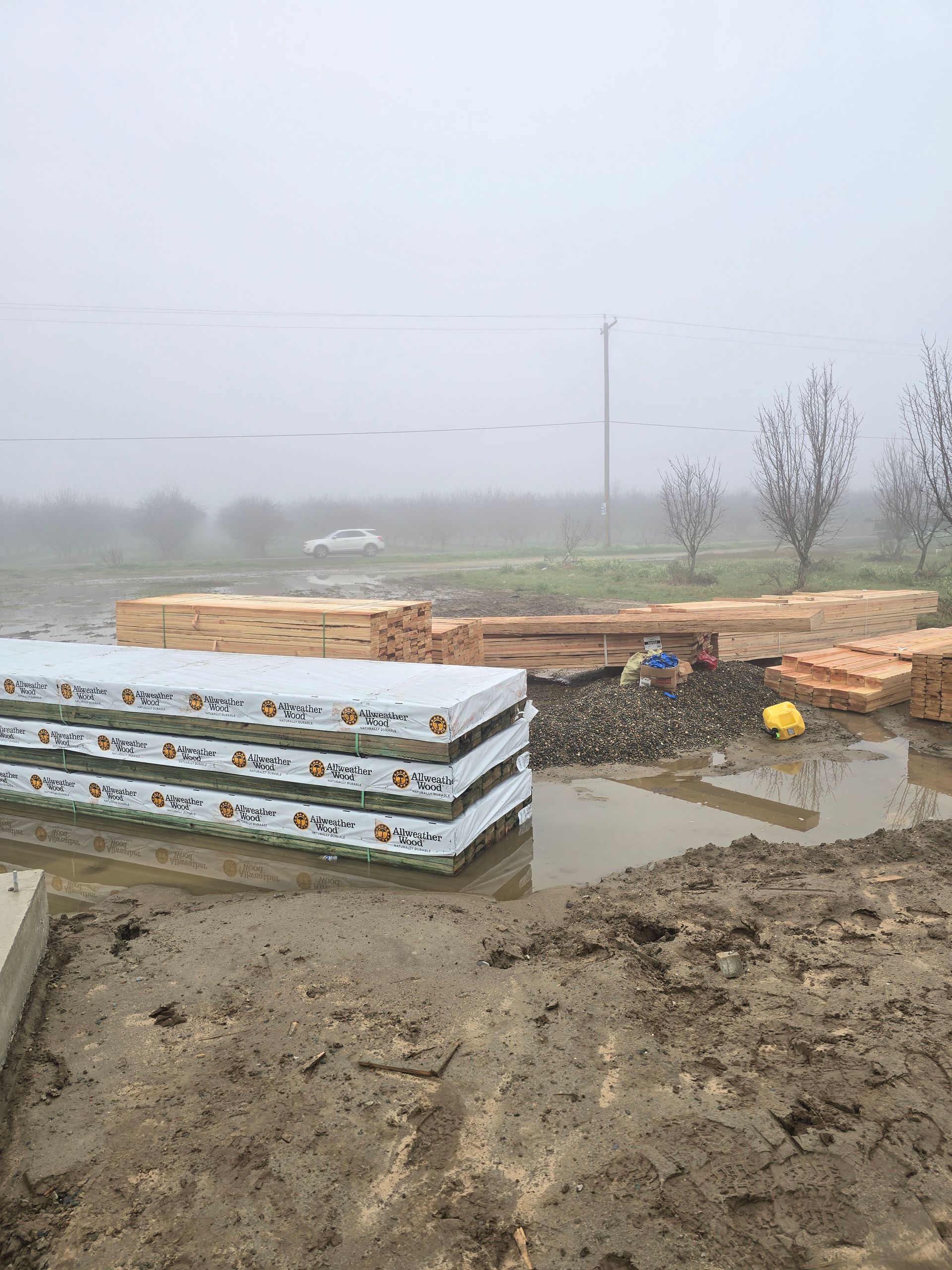 Piles of building materials and muddy ground at a foggy construction site near a roadside