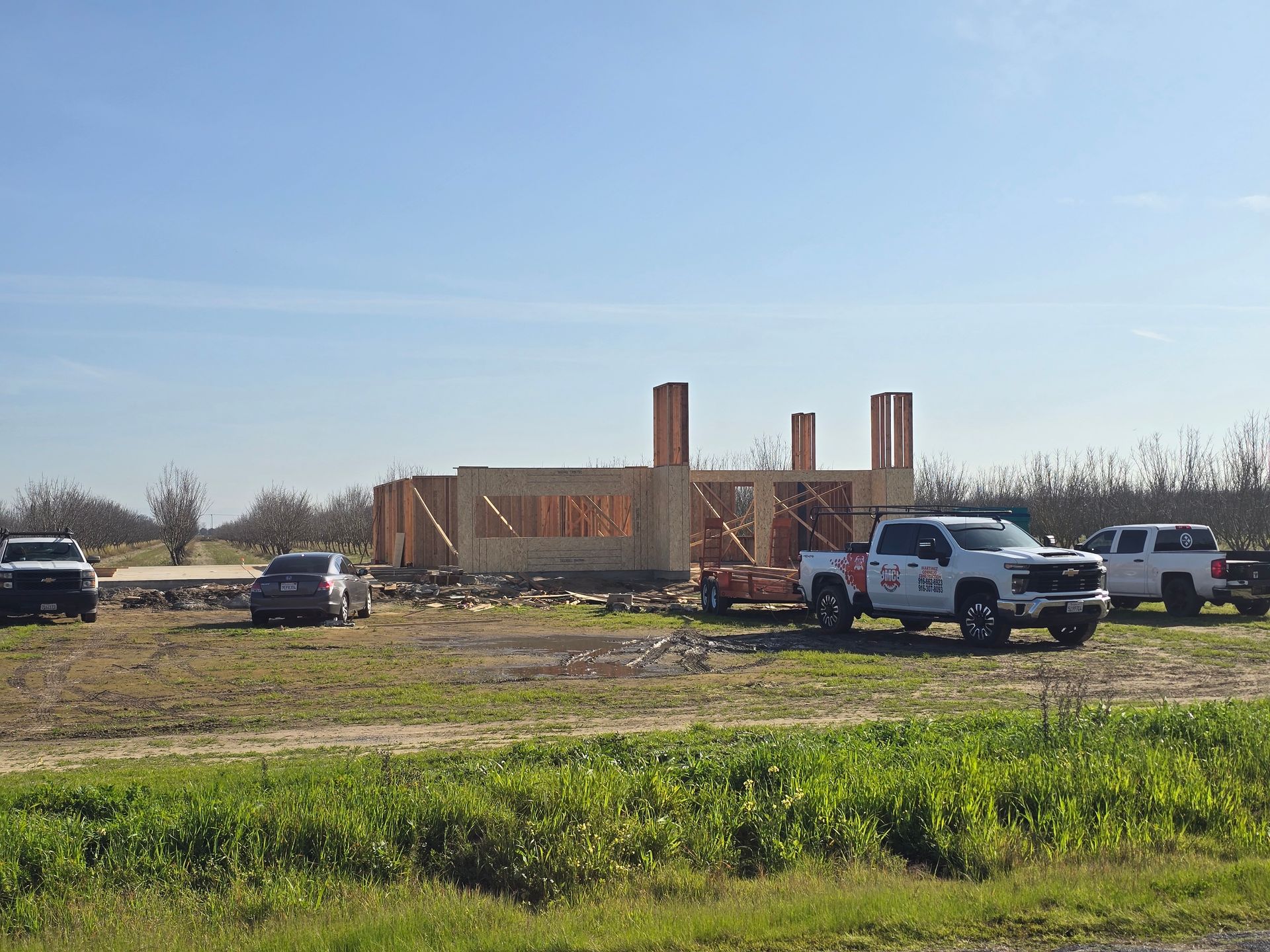 Construction site with brick walls rising in a field, surrounded by parked trucks under a clear sky