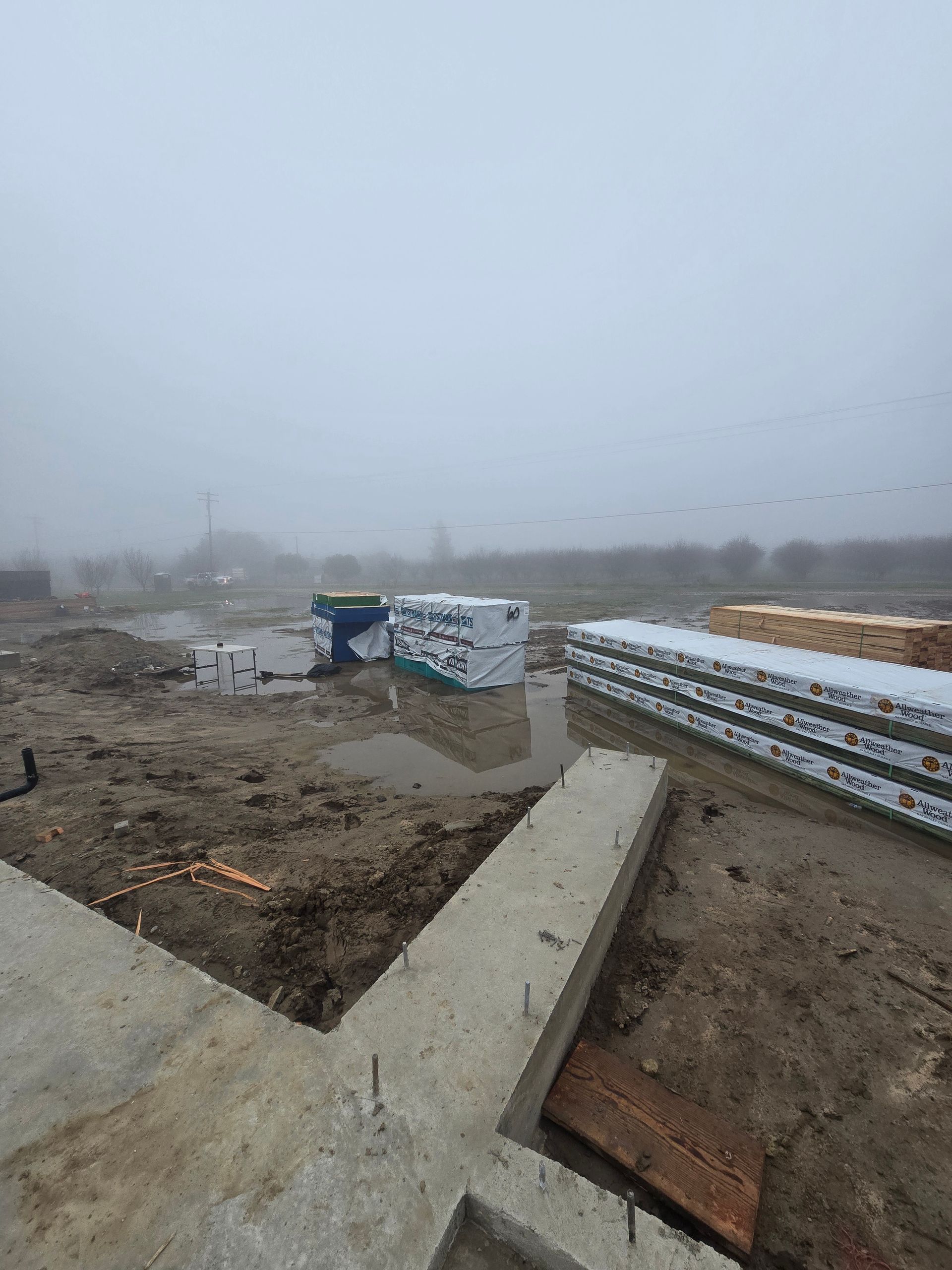 Foggy construction site with concrete slabs, dirt trenches, and stacked materials under a gray sky