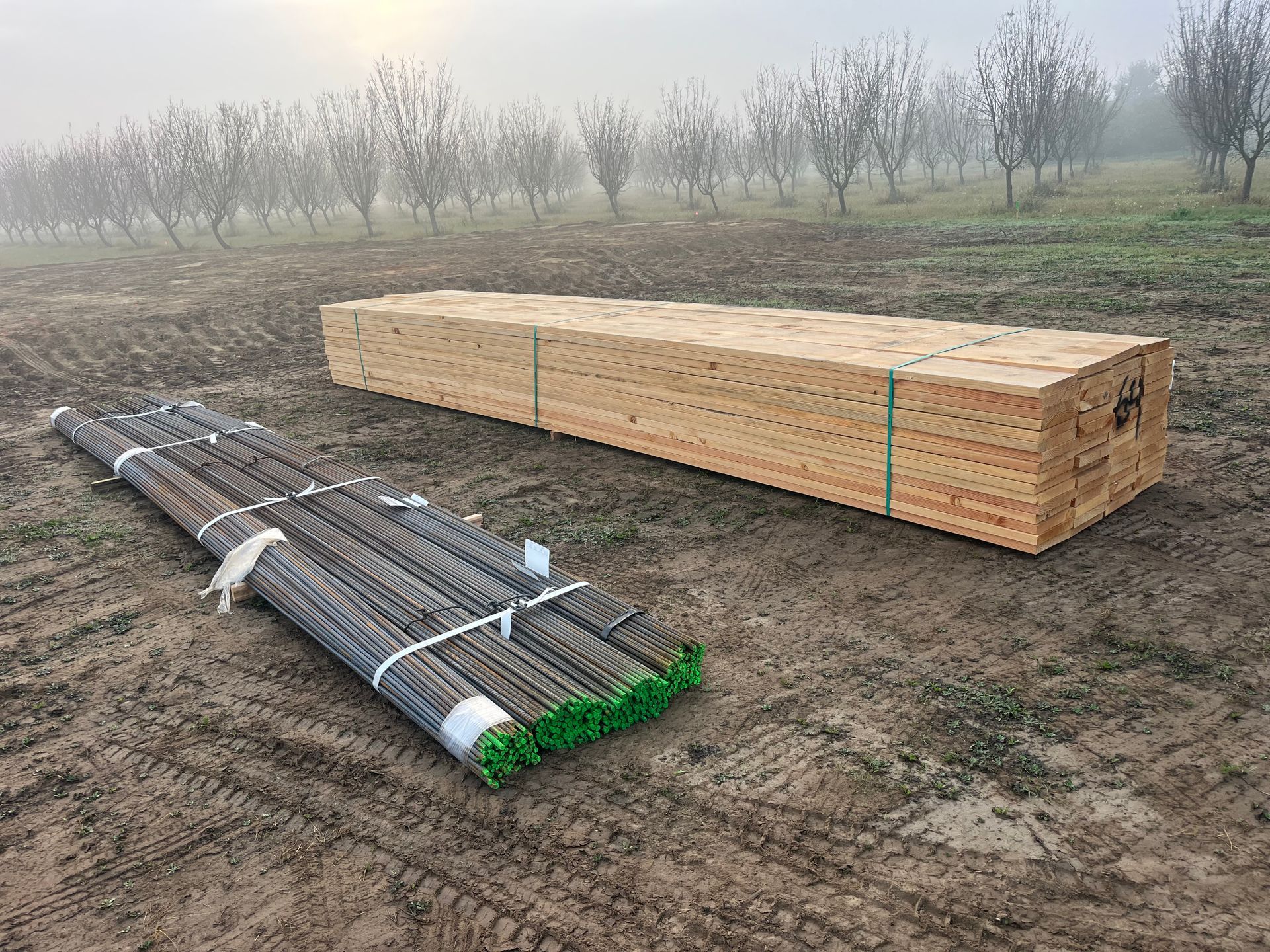 Bundles of wooden lumber and wrapped metal rods lying on a muddy field with trees in the foggy background