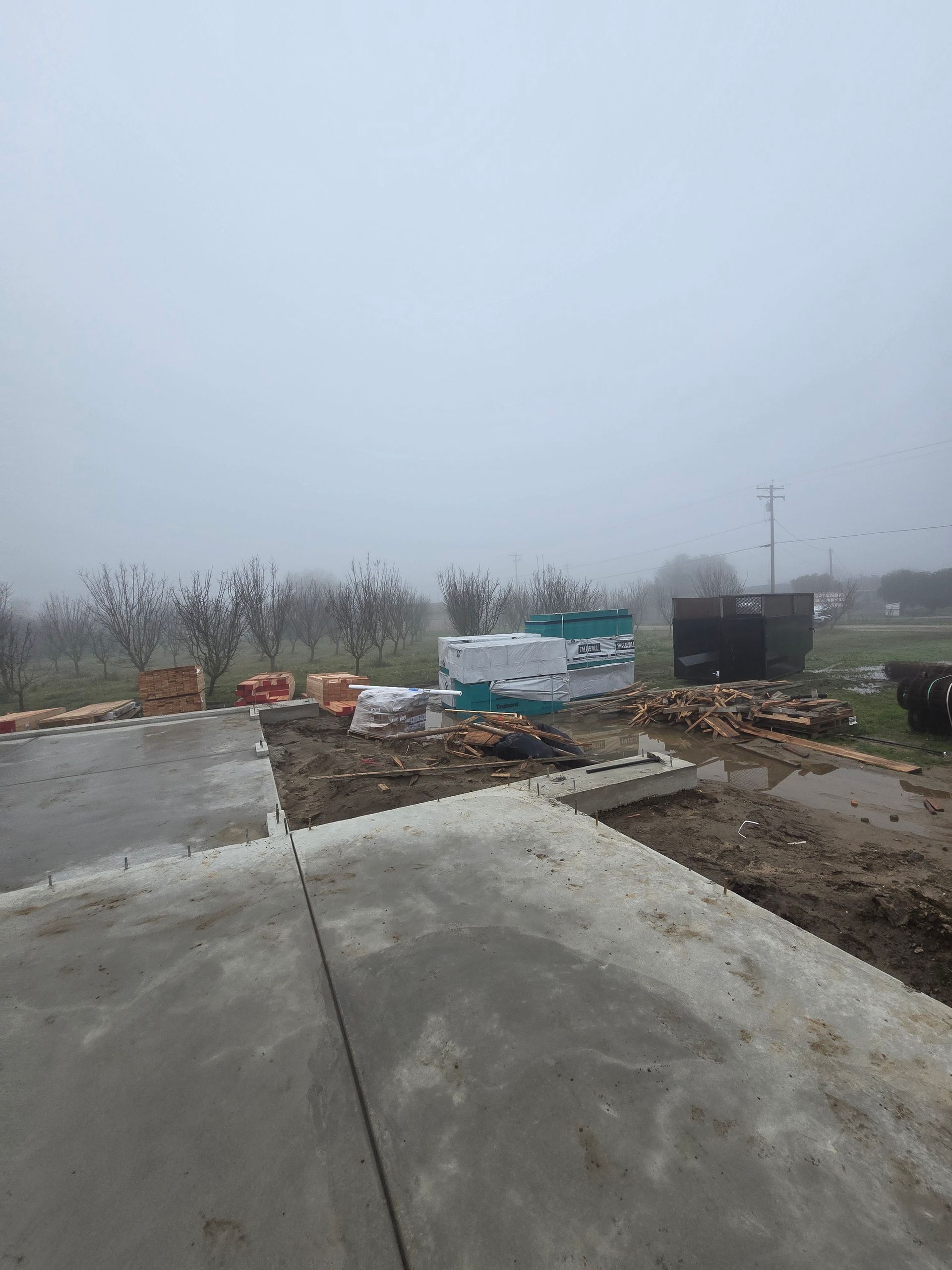 Foggy construction site with a wet concrete slab, dirt piles, barriers, and equipment in the background