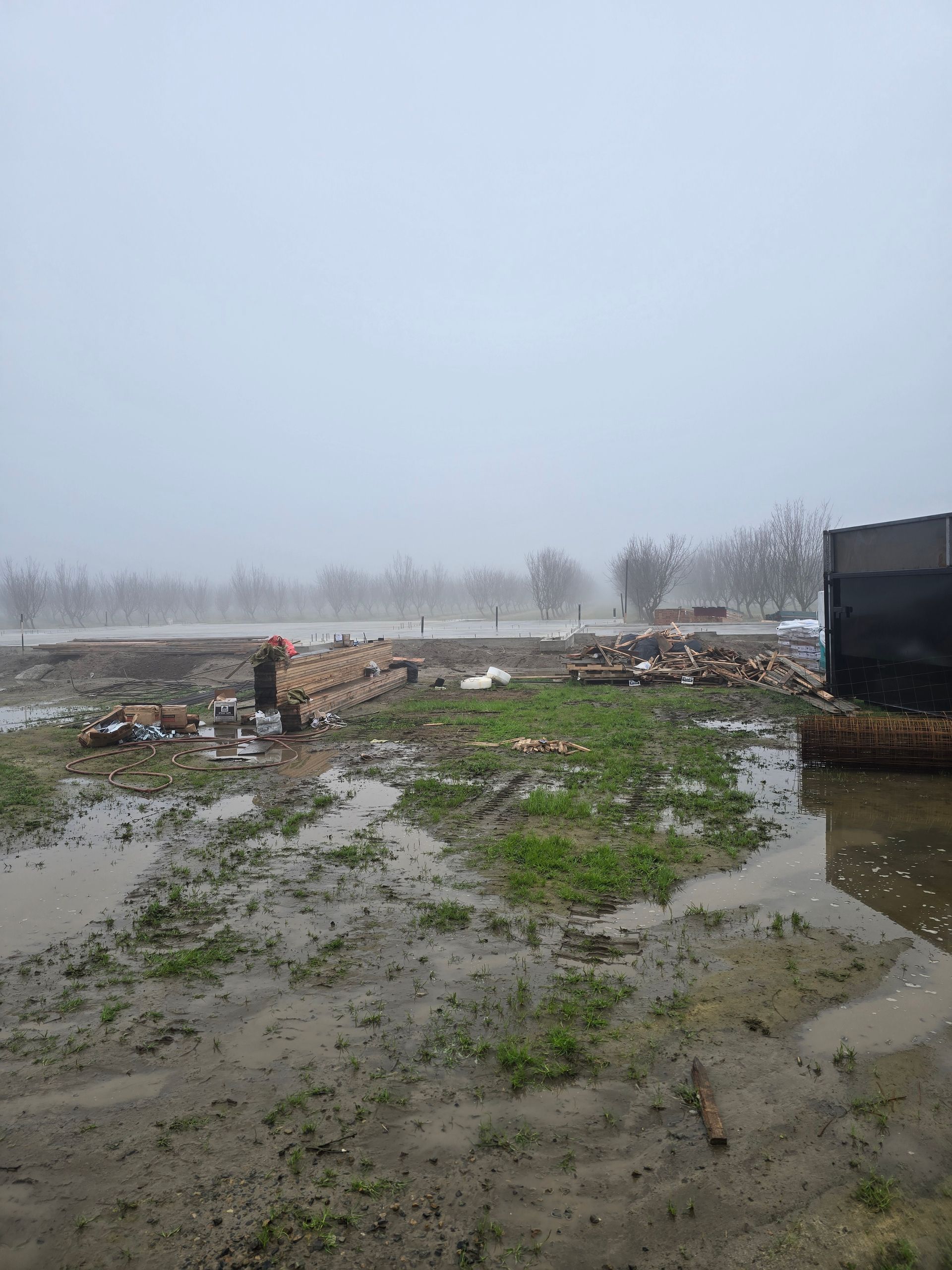 Flooded muddy field with scattered debris and a dark structure at right under an overcast sky