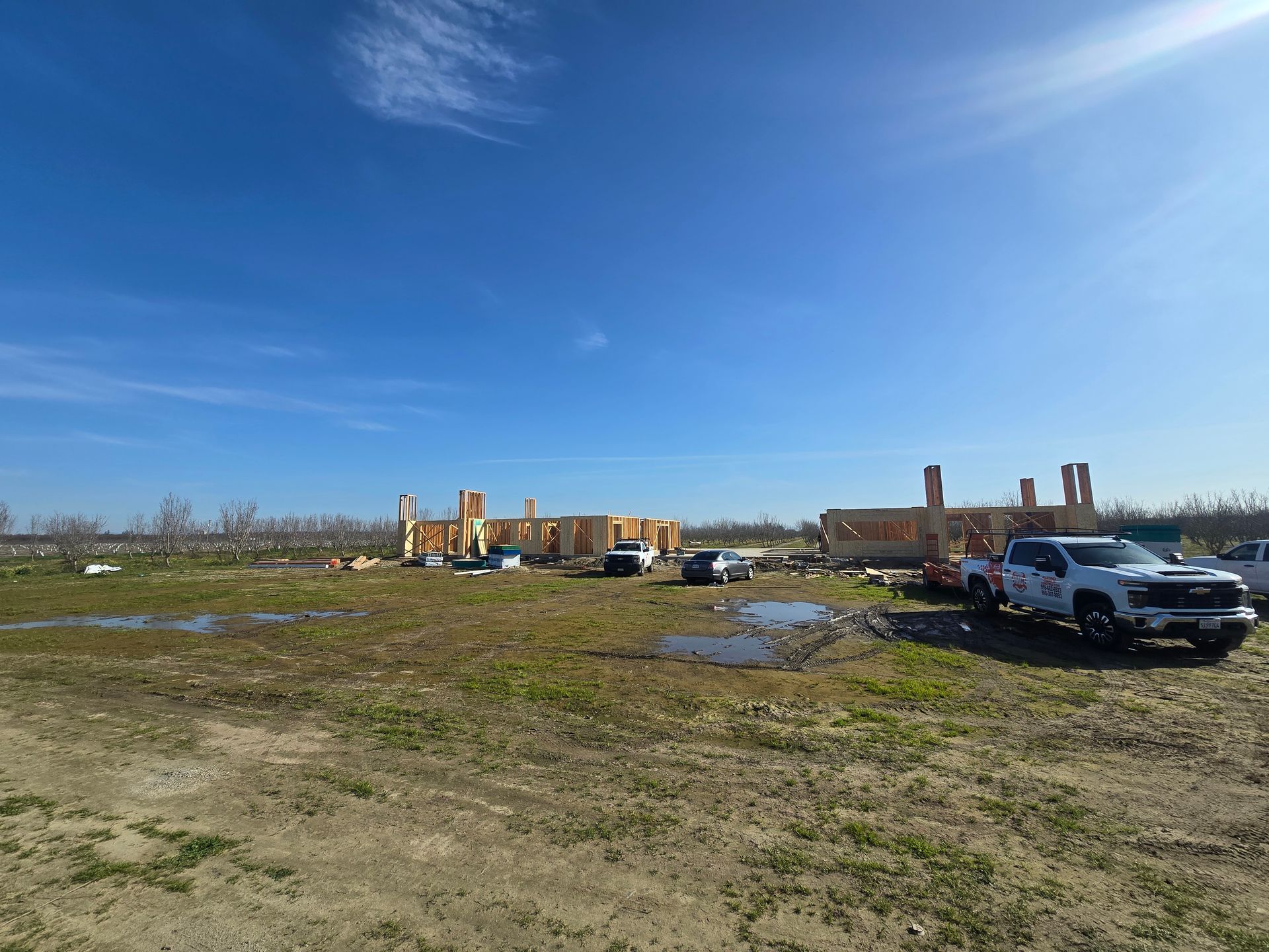 Open field with puddles, parked trucks, and a partly built brick structure under a blue sky.