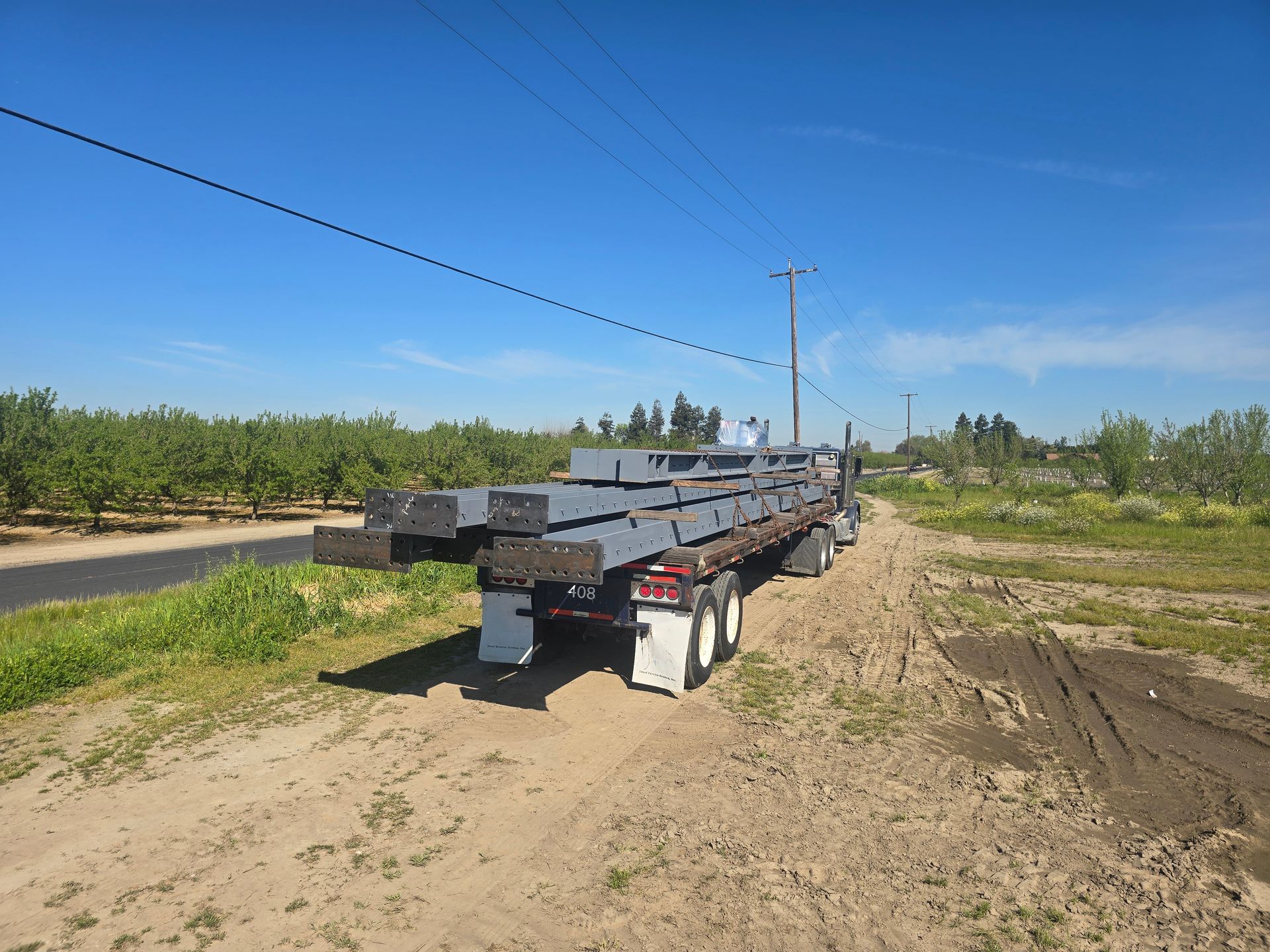 Flatbed trailer loaded with long metal beams on a dirt road beside orchard rows under a blue sky