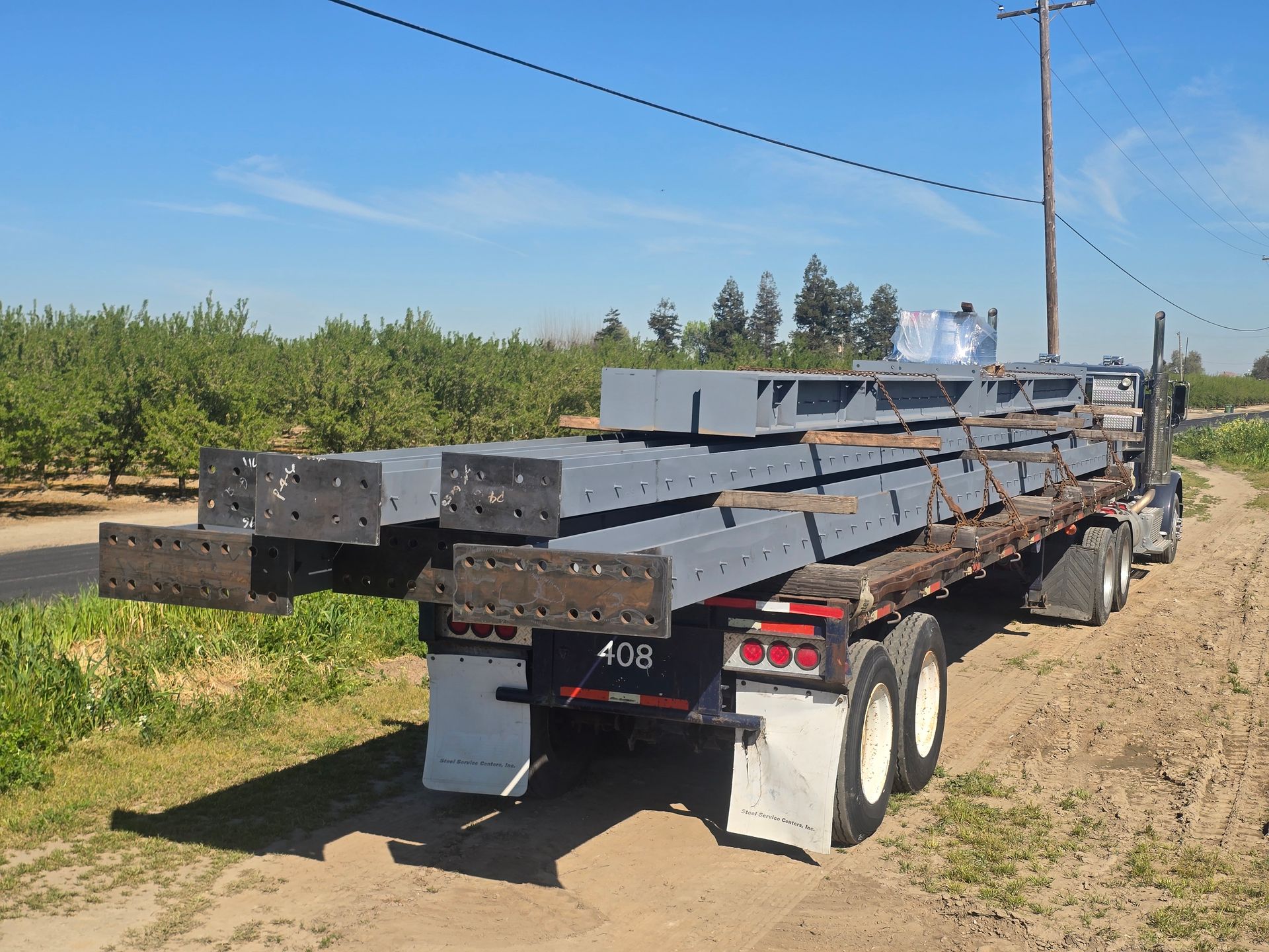 Flatbed trailer loaded with long dark steel beams on a dirt road beside orchards and blue sky