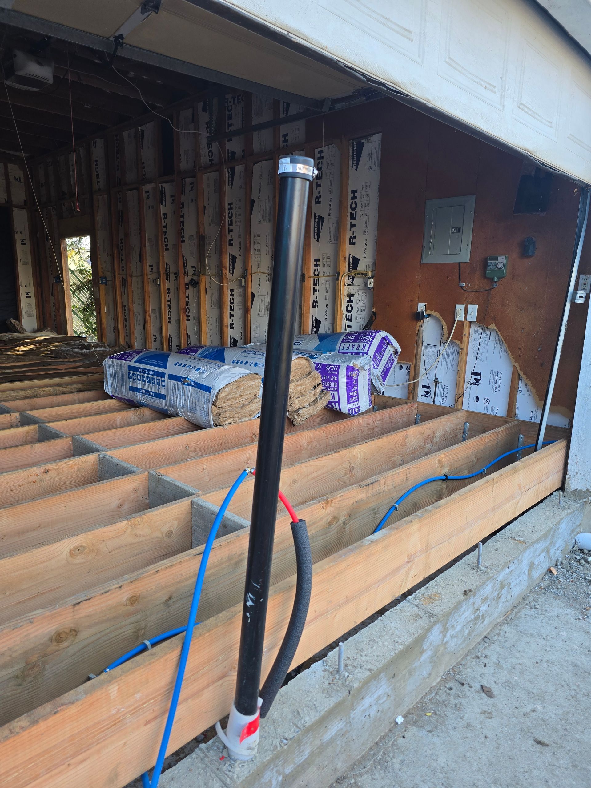 Construction area with exposed floor joists, wrapped insulation, and a black pipe in a basement opening