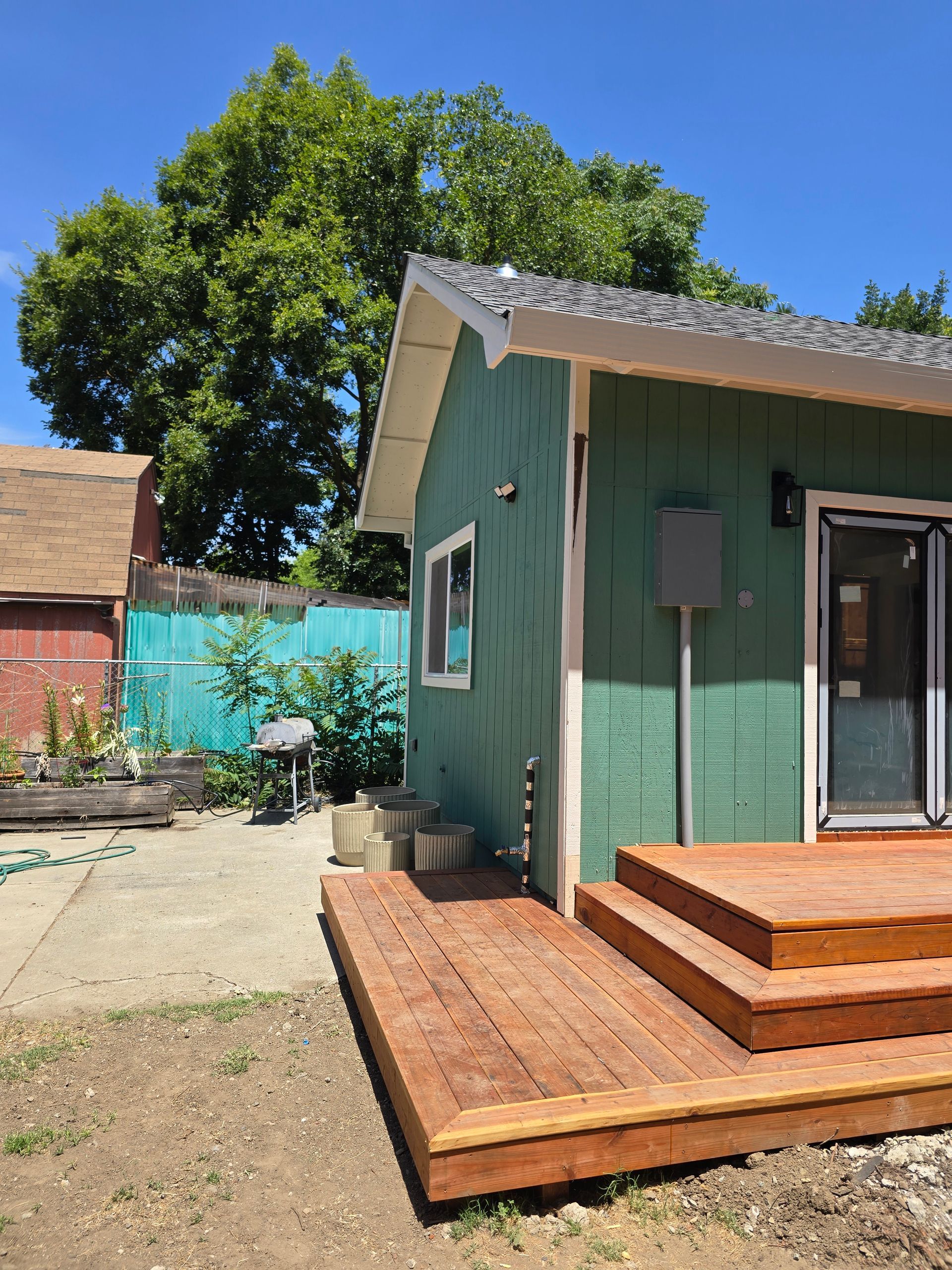 Green house with wooden deck and steps in a sunny yard, with trees and a red shed in the background