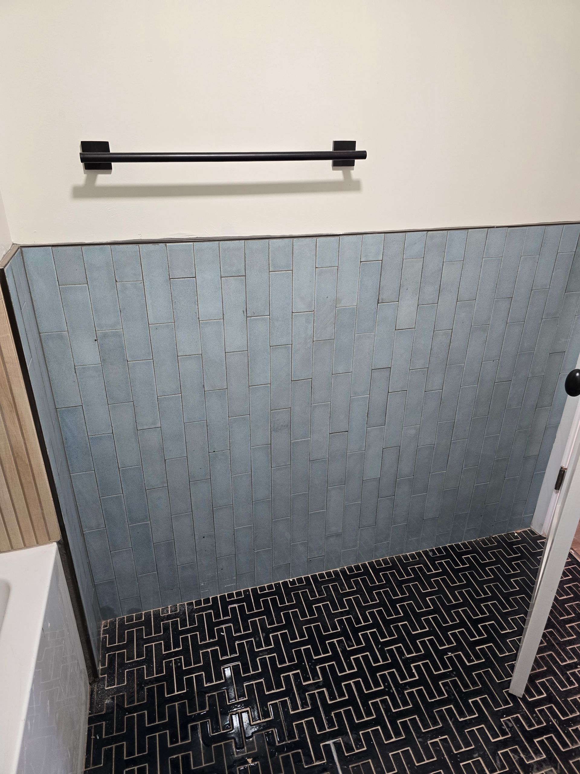 Empty tiled shower corner with a wall-mounted grab bar and black textured floor mat.