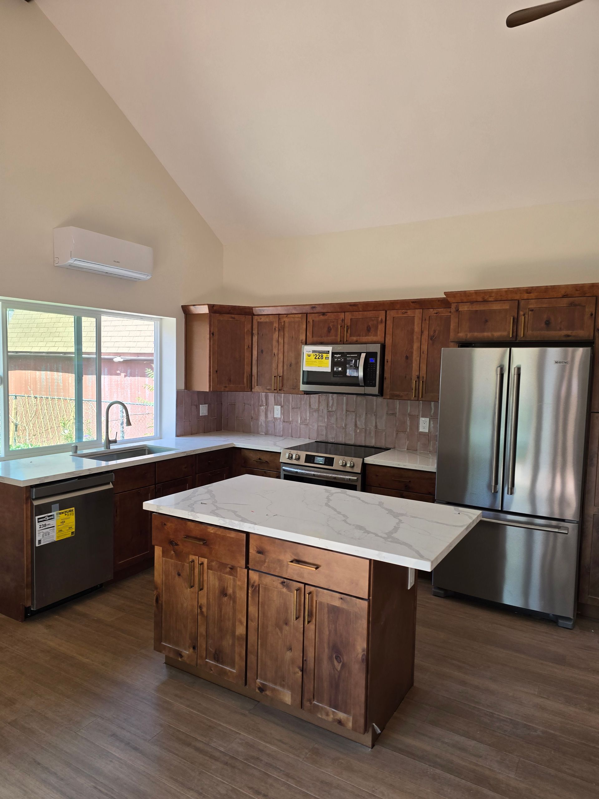 Empty kitchen with wood cabinets, stainless fridge, white island, and large window.