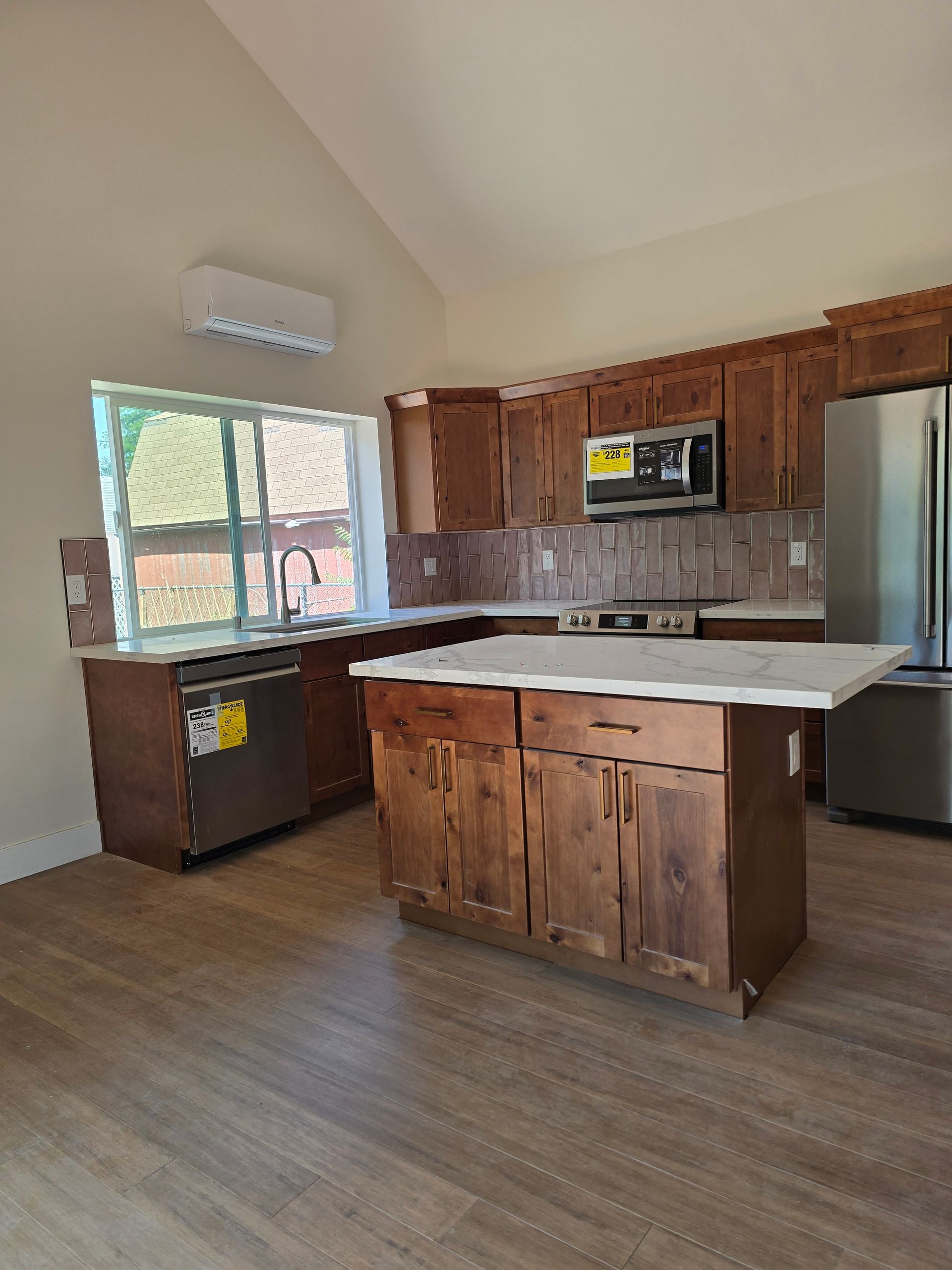 Empty kitchen with wooden cabinets, white island countertop, and large window letting in natural light.