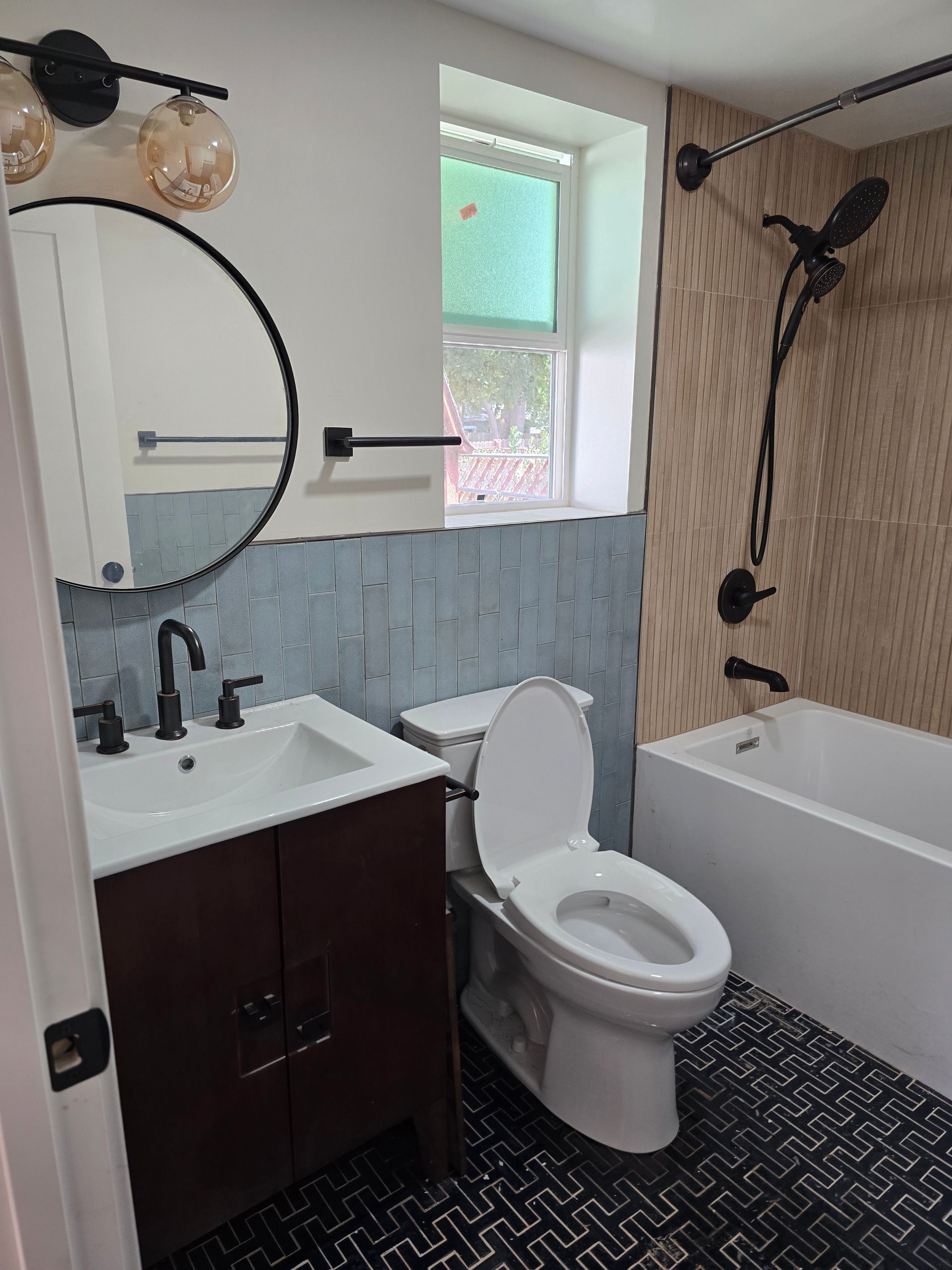 Small bathroom with white tub, toilet, vanity, round mirror, and patterned wallpaper accents.