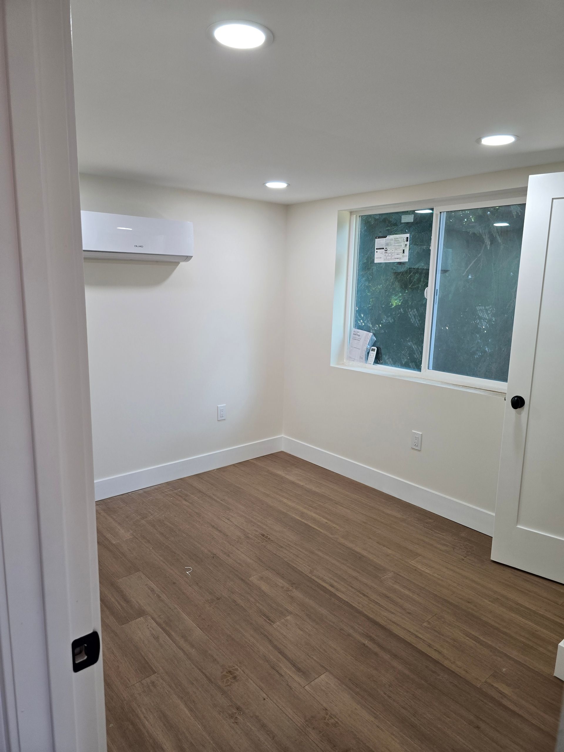 Empty white room with wood-look floor, recessed lights, and a window with plastic covering