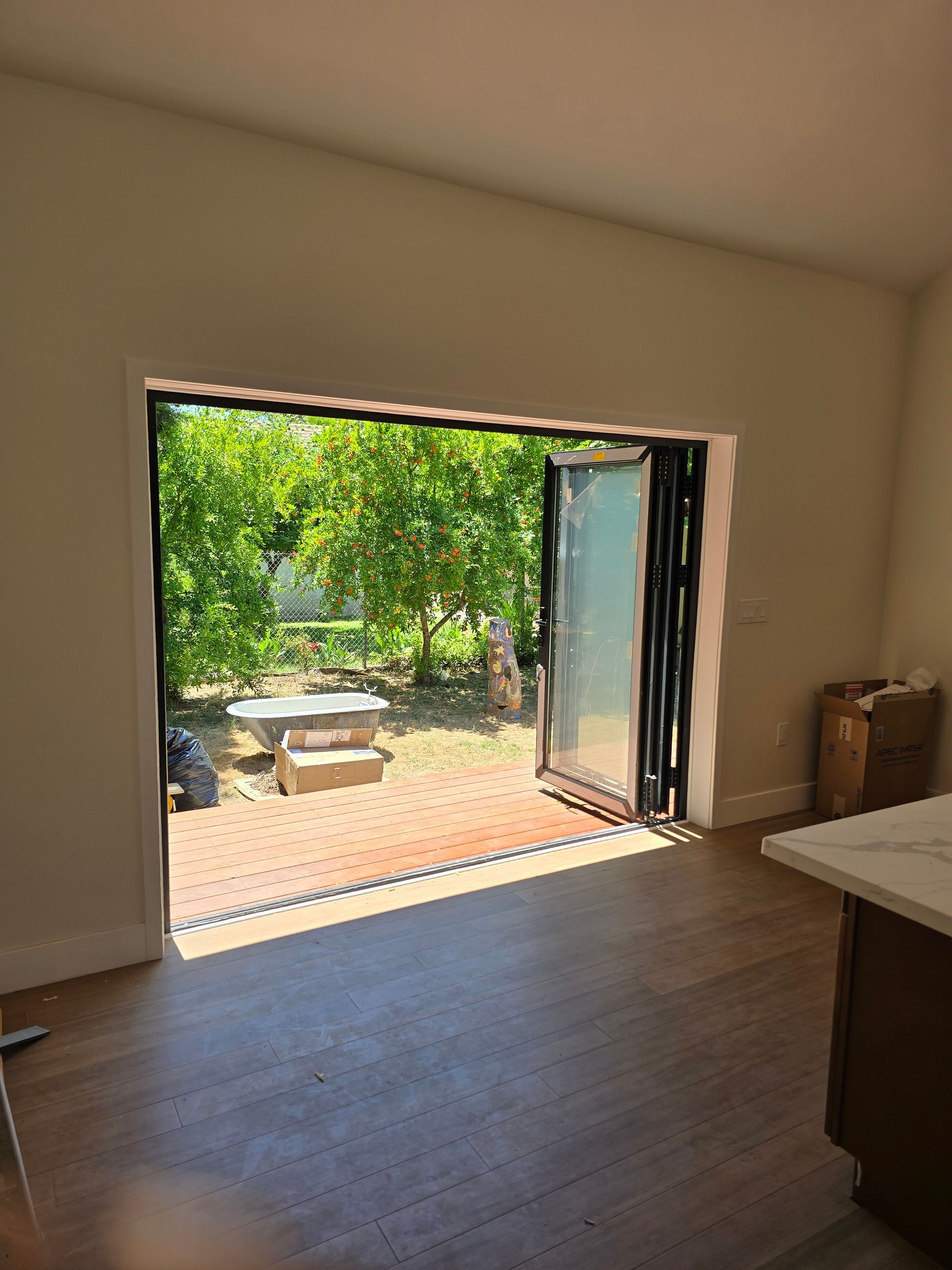 Open room with wood floor and wide doorway to sunny patio with table and green trees