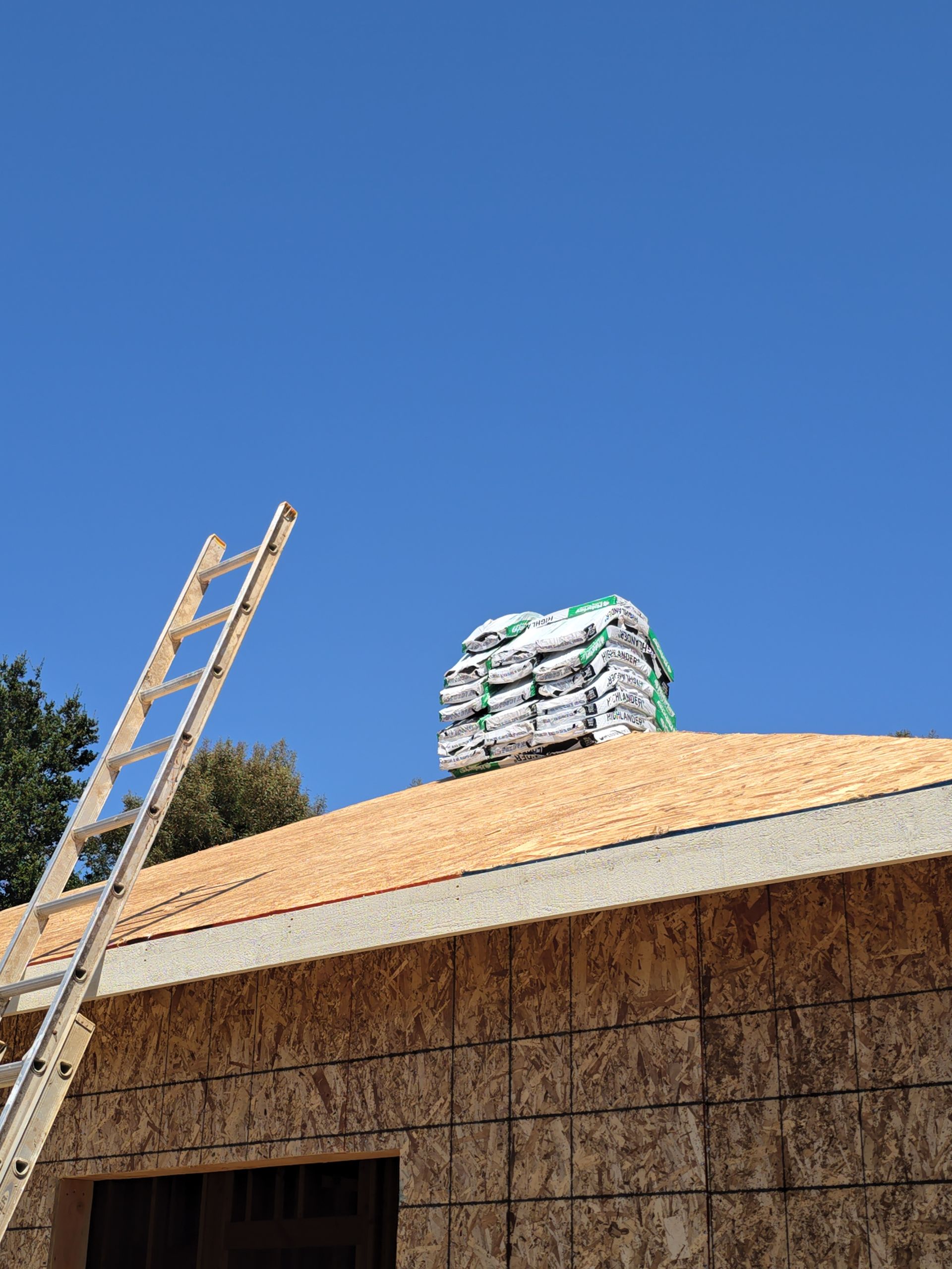 Ladder leaning against a roof under construction with stacked shingles on top against a blue sky