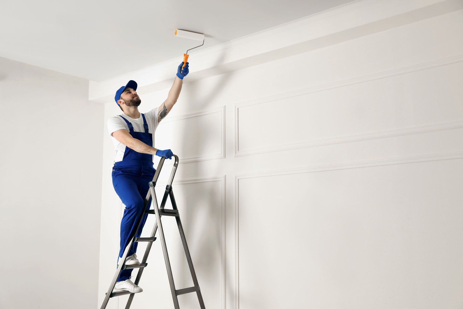 Painter in blue overalls painting a white ceiling from a ladder indoors