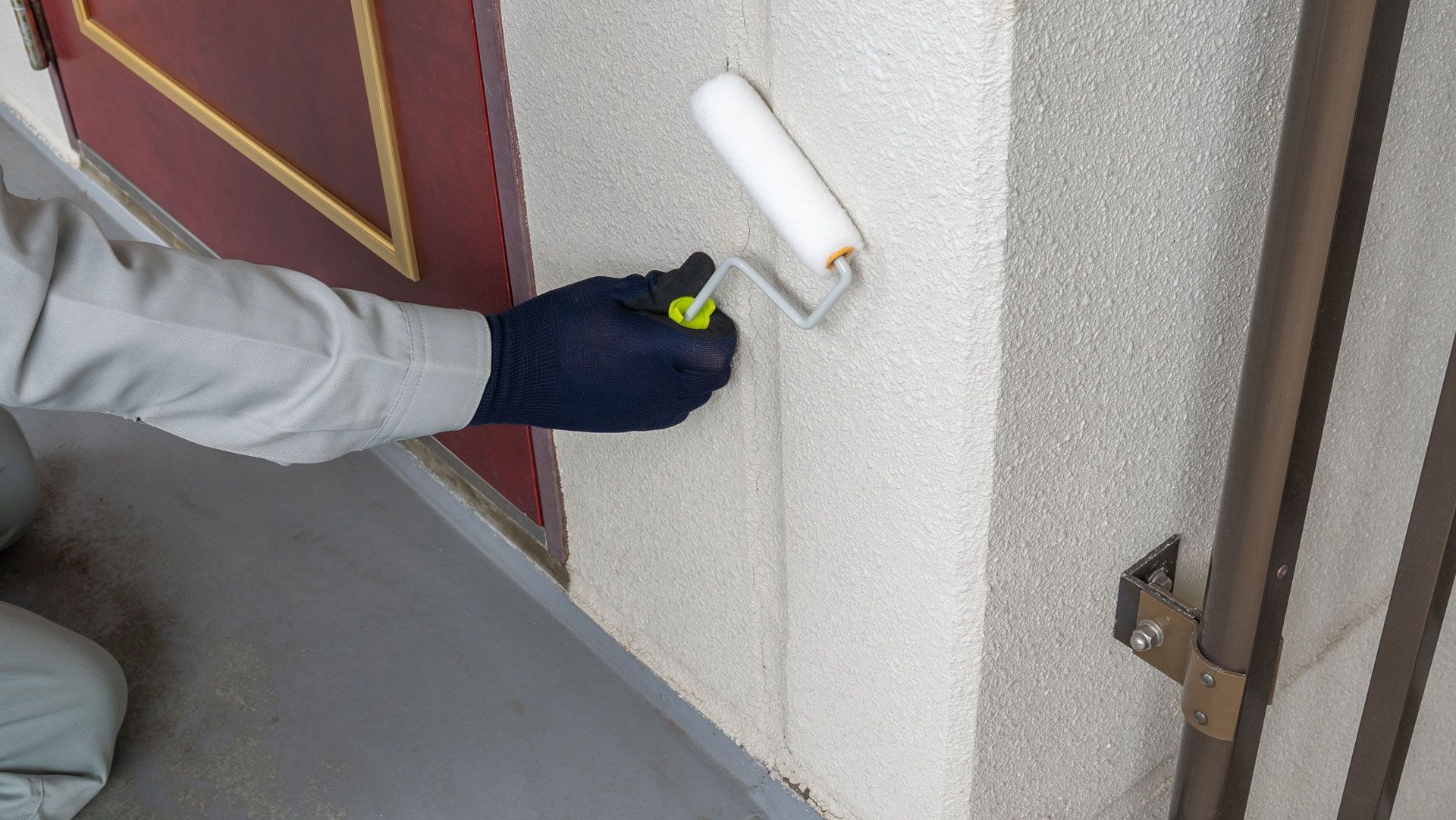 Gloved hand using a paint roller to coat a light-colored wall beside a brown door.
