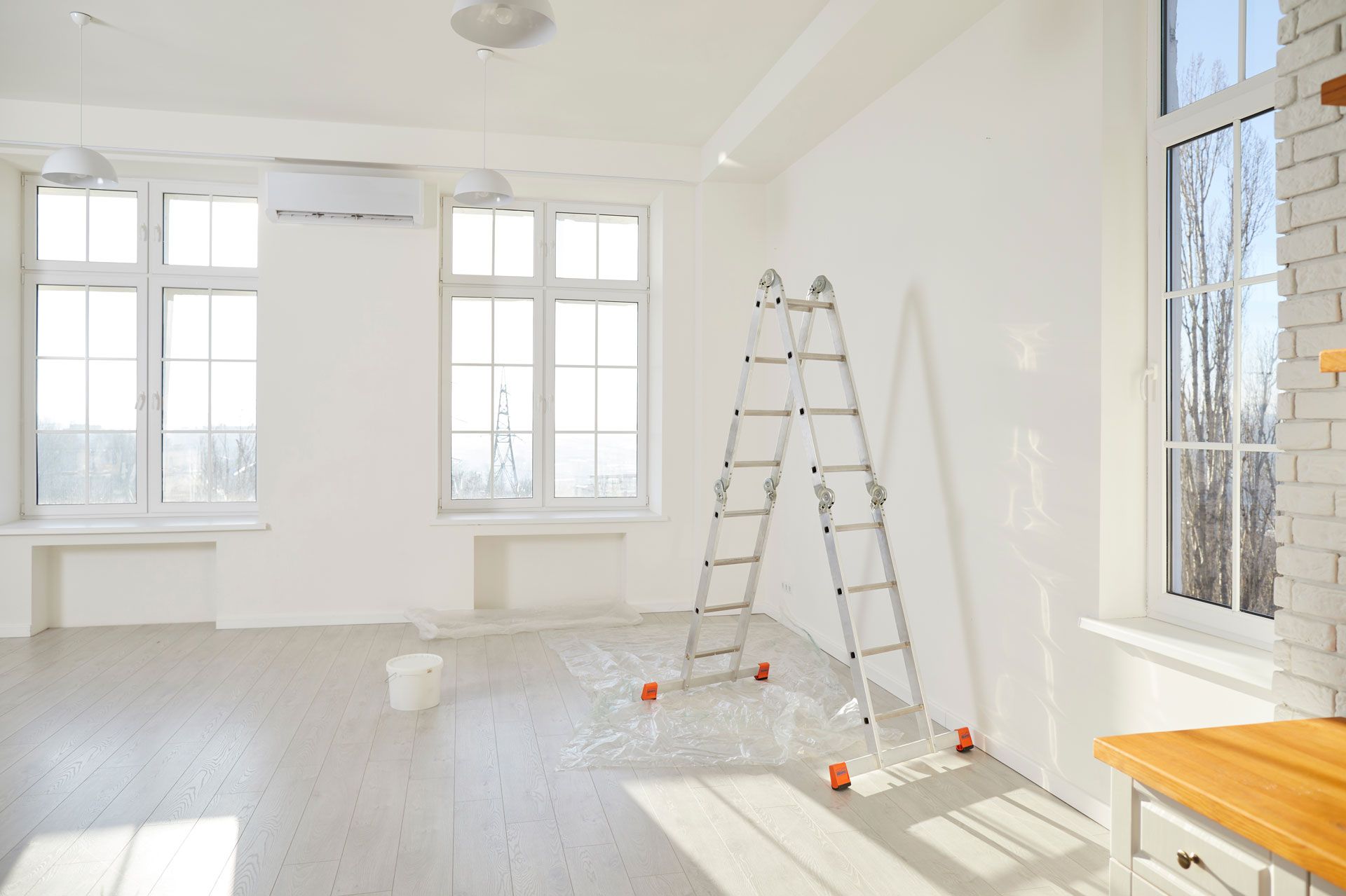 Bright empty room with white walls, large windows, and a step ladder on the floor during painting or renovation