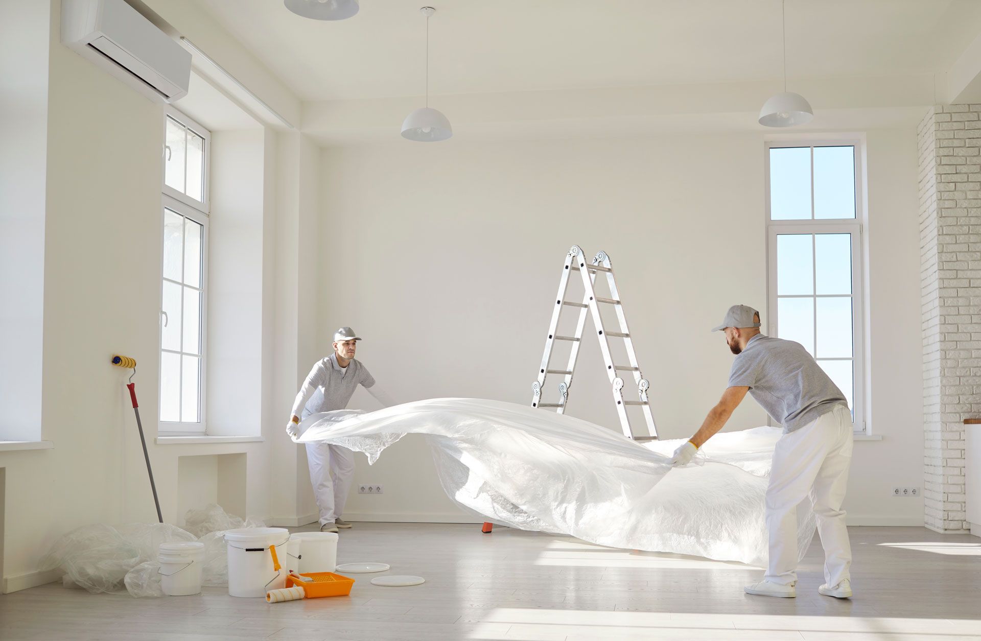 Two workers in white paint a bright room while covering furniture with plastic and a ladder stands nearby.