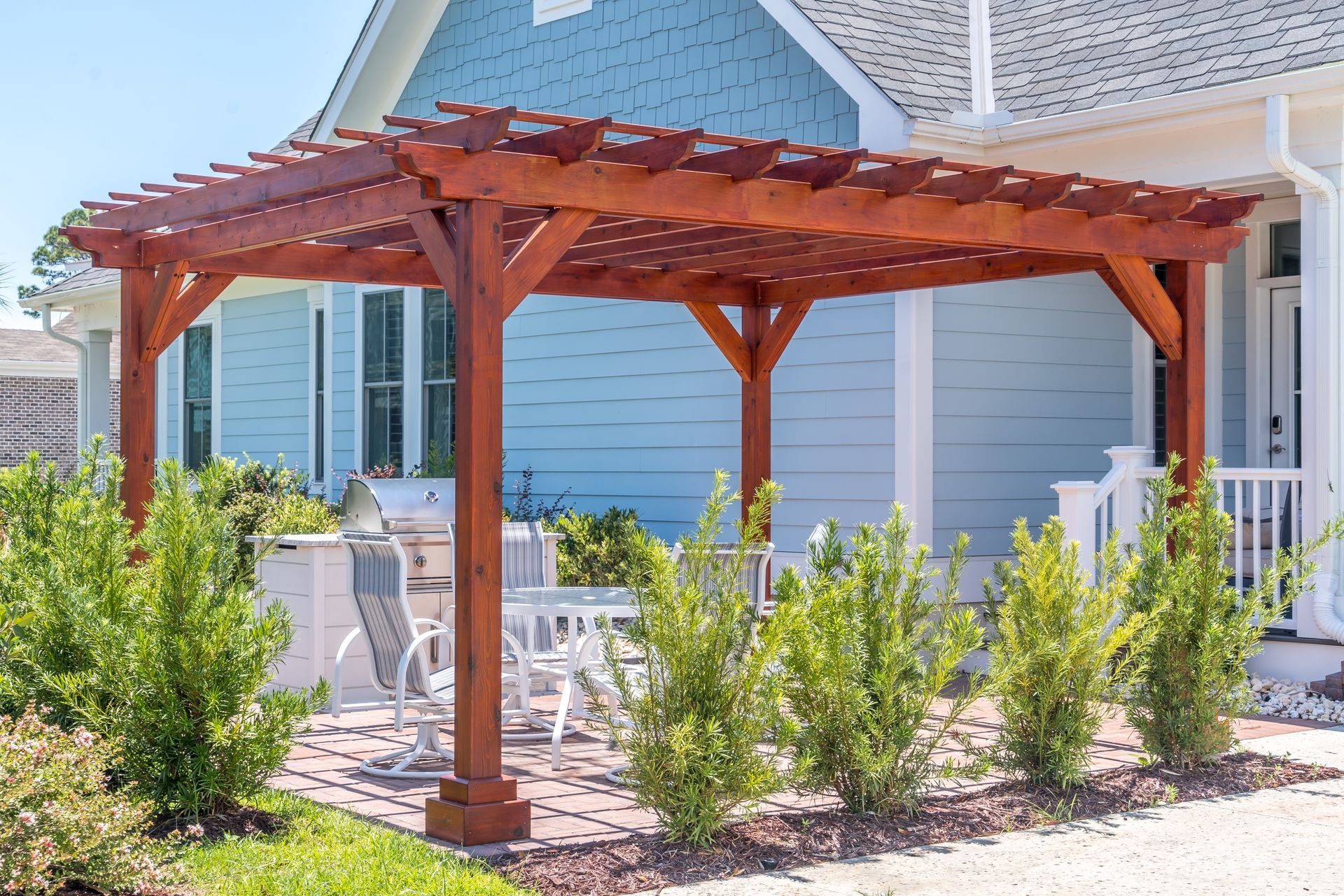 Wooden pergola with patio furniture beside a blue house and garden shrubs
