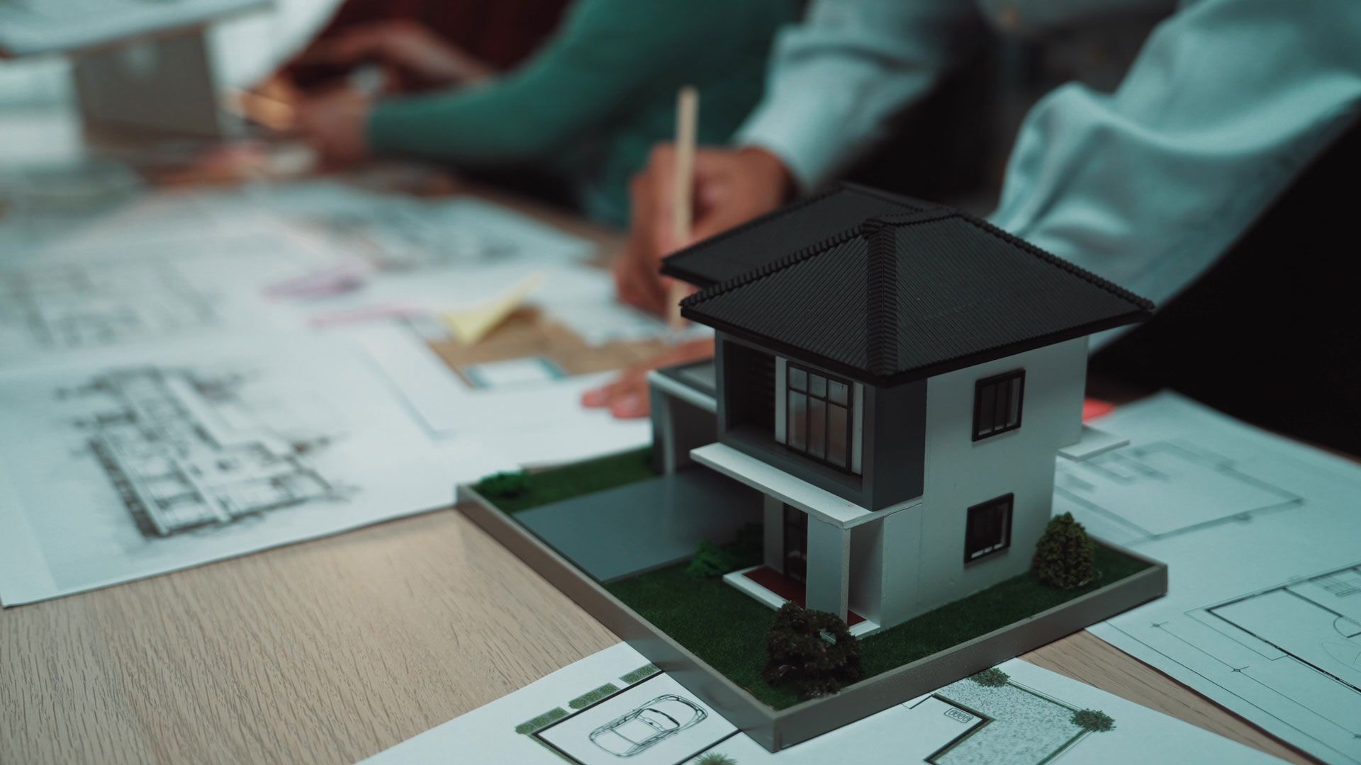 A small house model on architectural blueprints, with people reviewing plans in the background