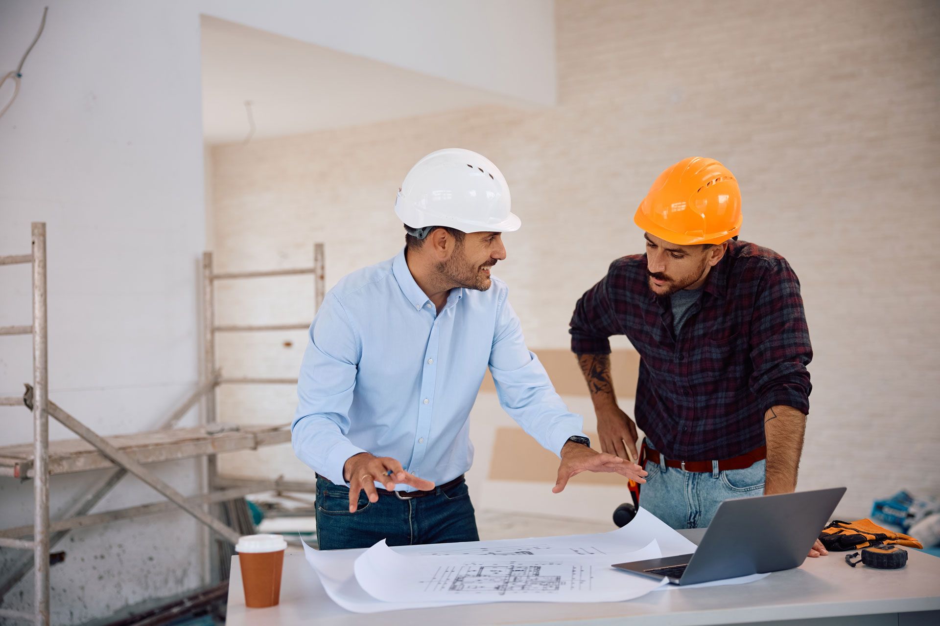 Two construction workers in hard hats reviewing blueprints at a table with a laptop and coffee cups
