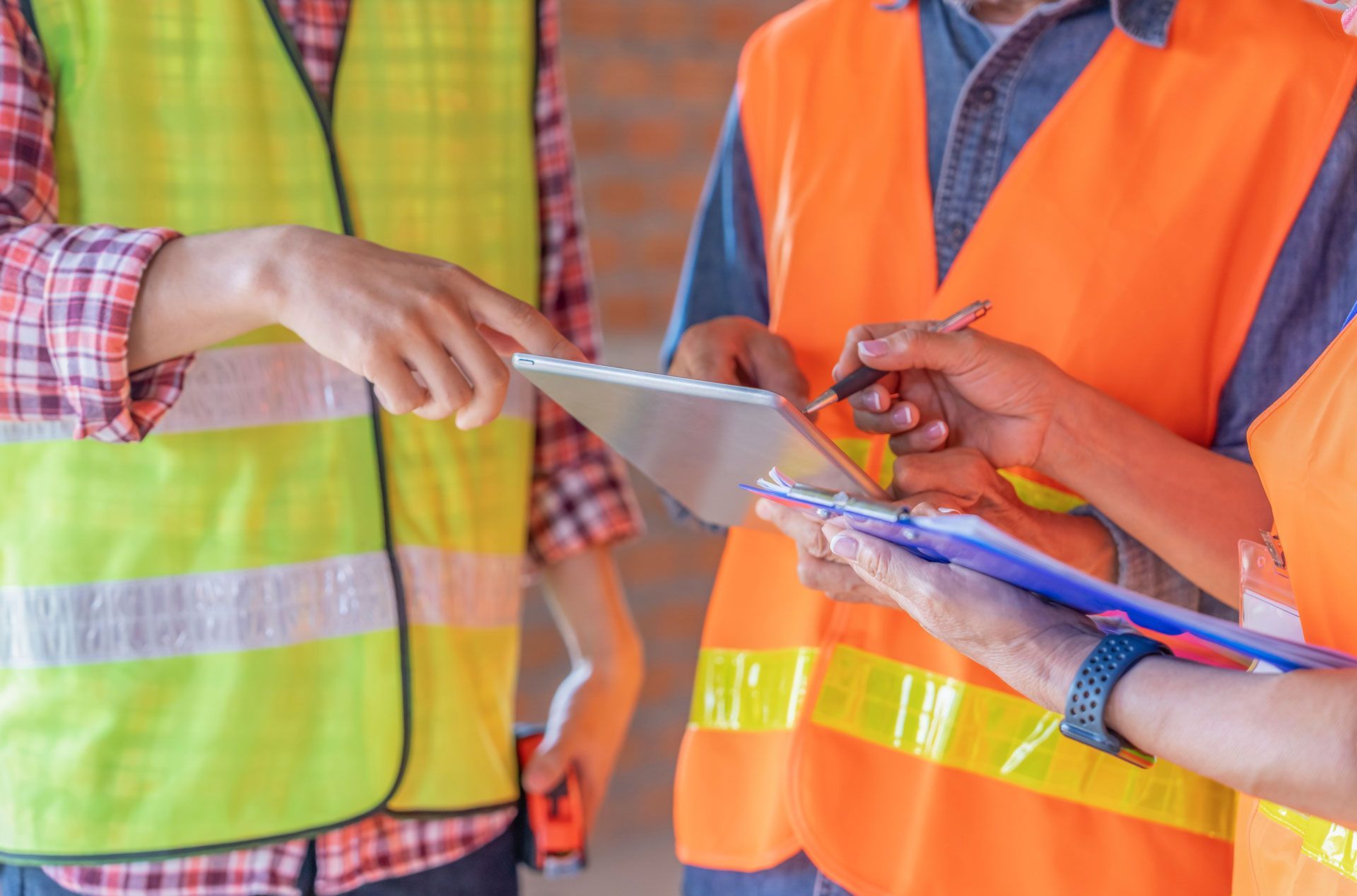 Workers in orange and yellow safety vests reviewing a tablet and clipboard on a construction site