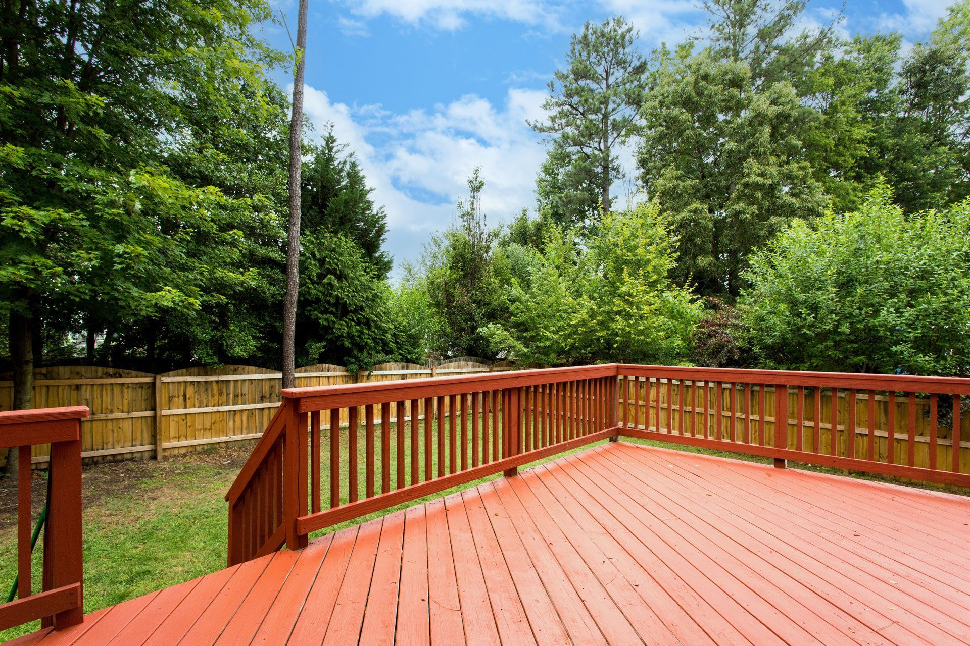 Red wooden deck with railing overlooking a green backyard and trees under a blue sky