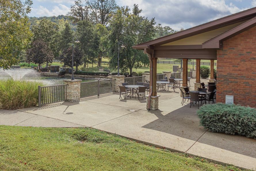 Brick patio beside a brick building, with tables, chairs, and a pond view beyond a grassy lawn.