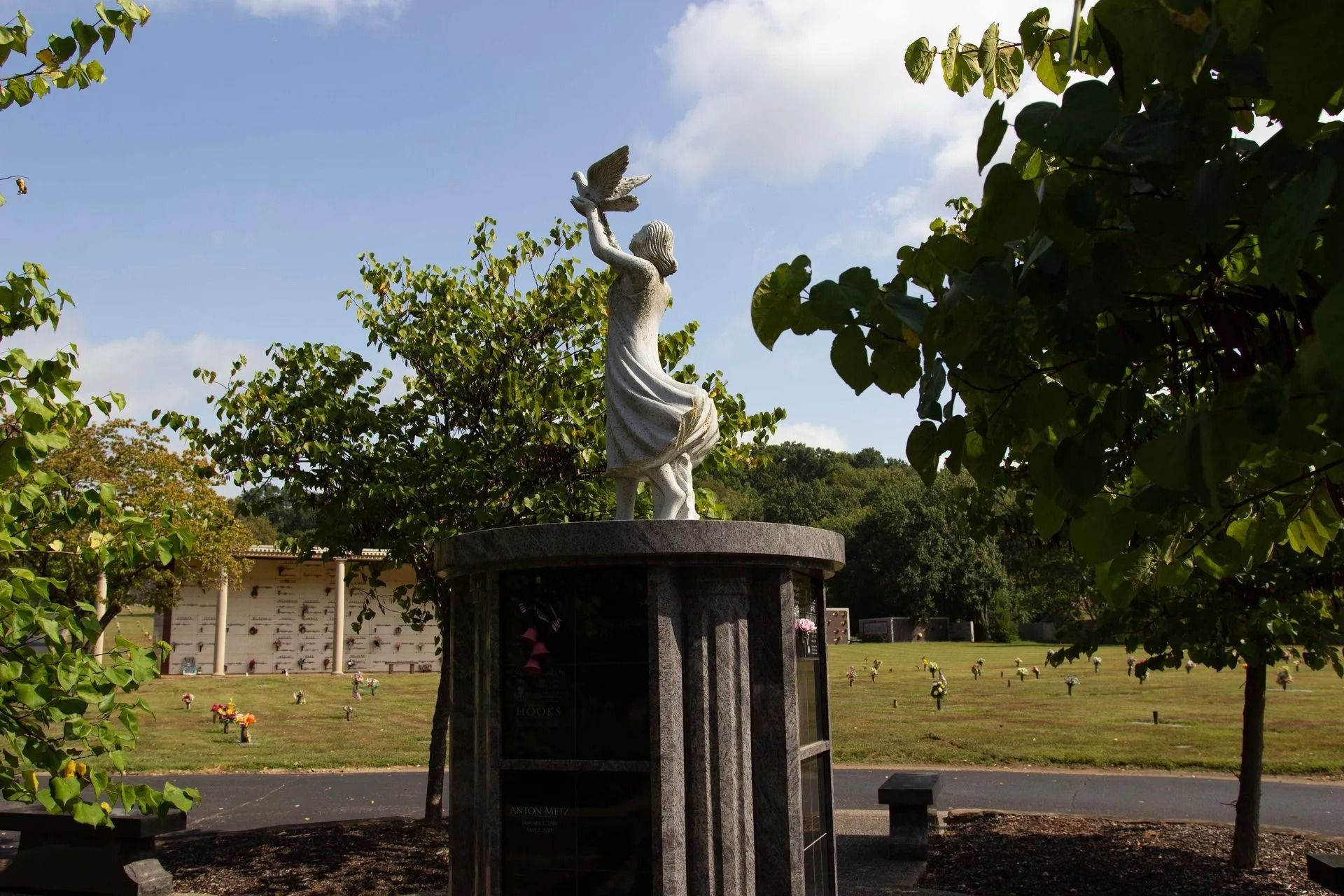 White statue on a pedestal in a sunny park with trees and a grassy field in the background