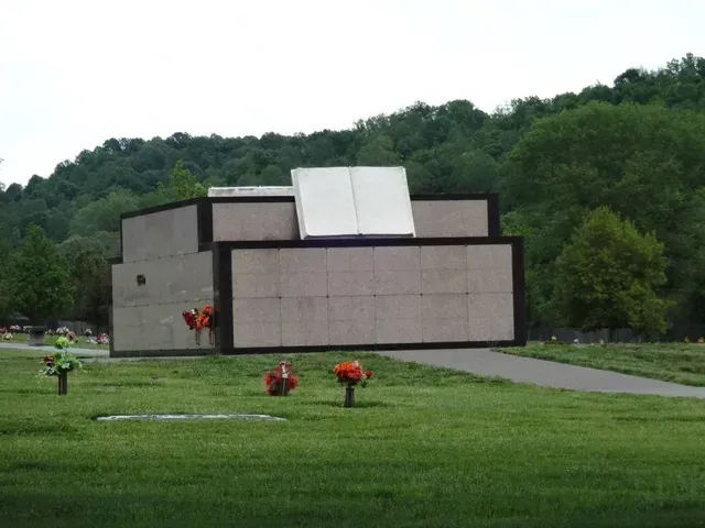 Modern abstract monument on a grassy lawn with flowers, backed by a wooded hillside