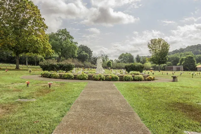 Path leading to a fountain in a green park with trees and cloudy sky